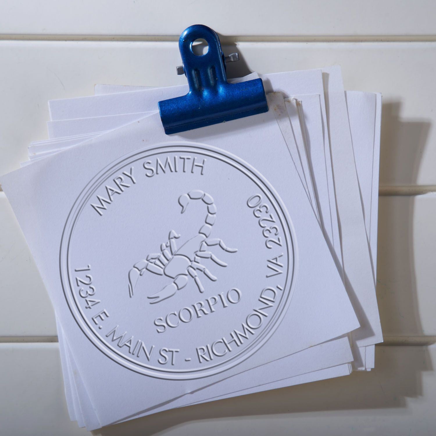 Blue binder clip holding white embossed stamp and seal samples with scorpion seal, Mary Smith name, and Richmond address.