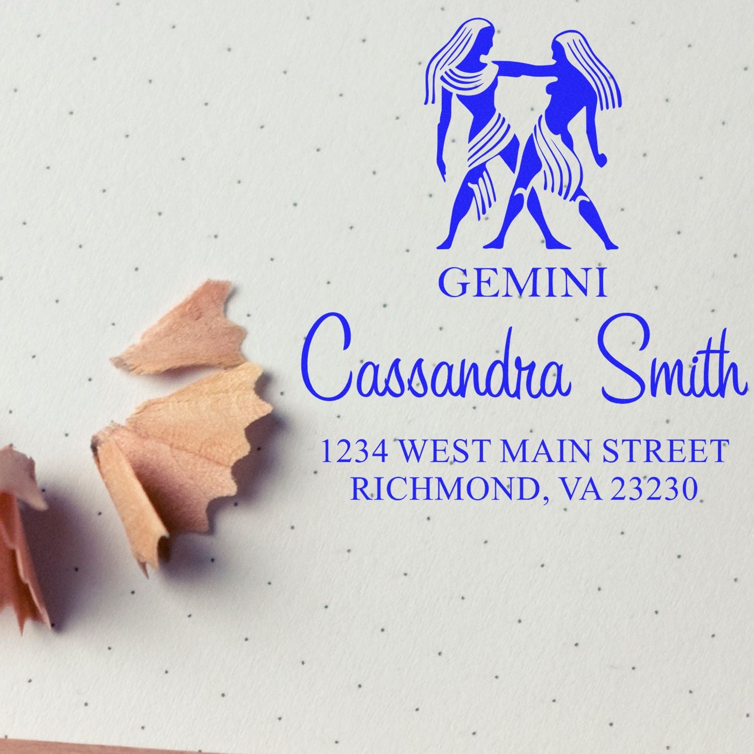 Blue Gemini custom stamp seal with twin figures, Cassandra Smith name, and Richmond address on dotted paper with pencil shavings.