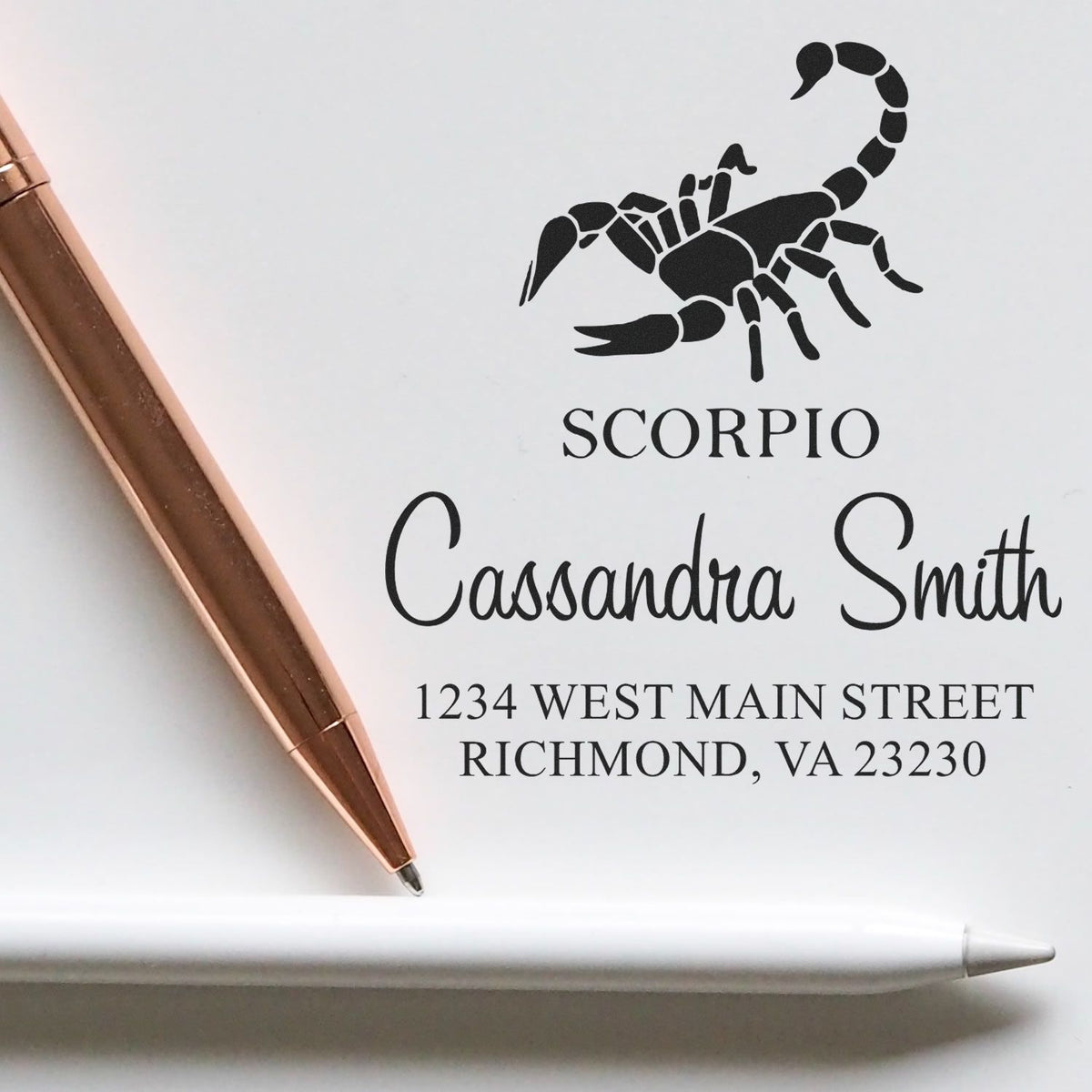 White custom address stamp with black scorpion seal, Scorpio name and address, beside a rose gold pen and white stylus.