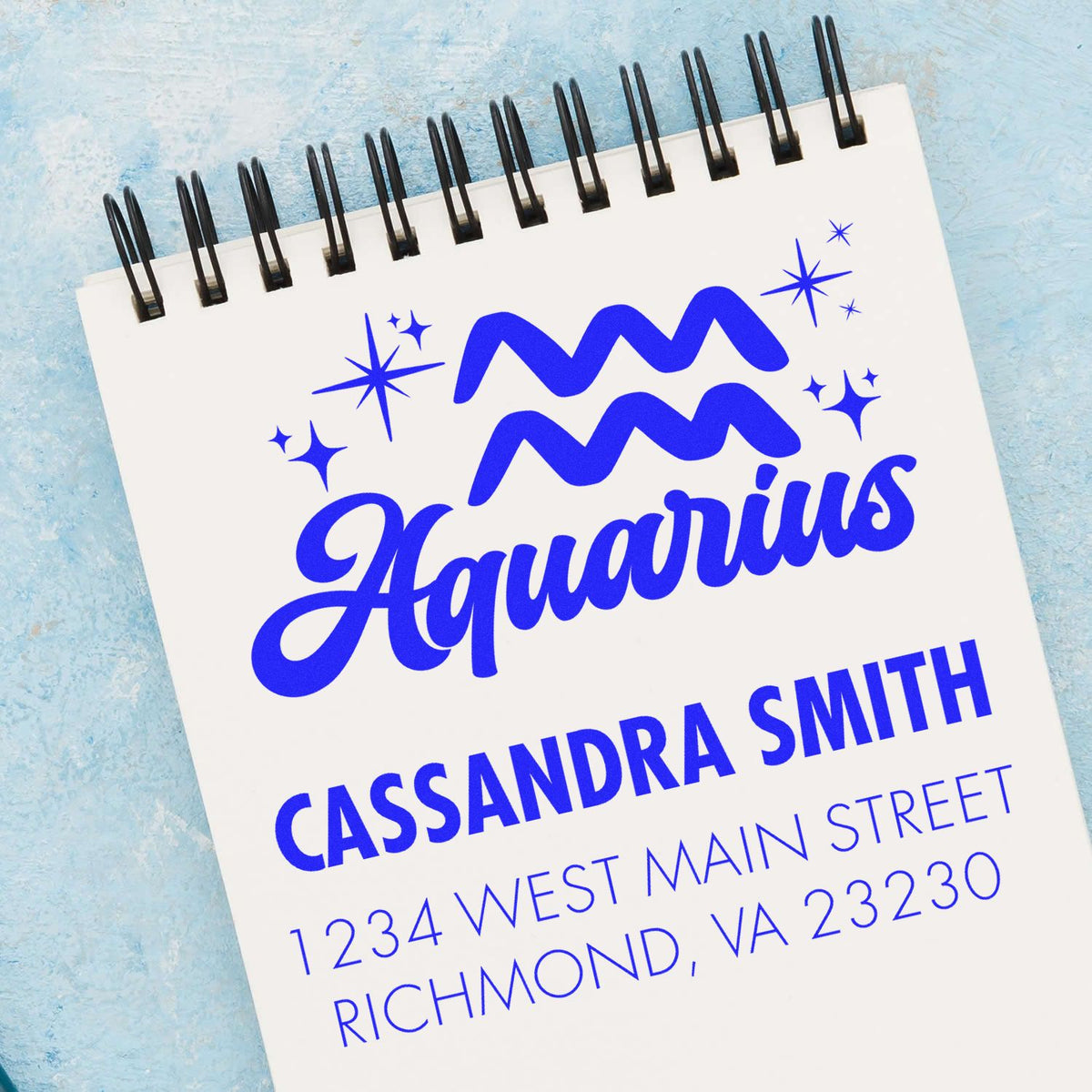 Blue Aquarius personalized stamp and seal design on white spiral notebook with black binding, stars, and Cassandra Smith address.