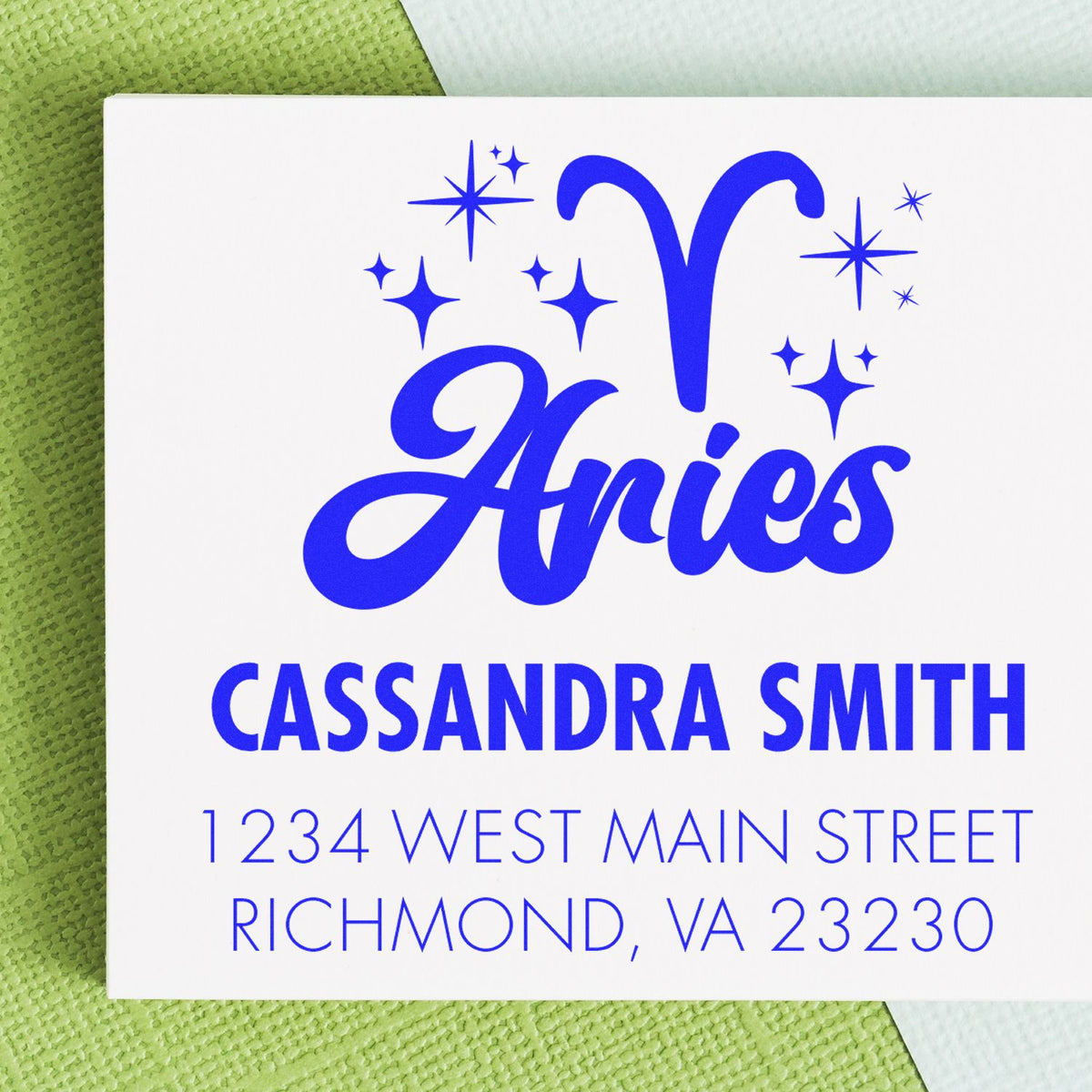 Blue Aries custom stamp and seal in bold cobalt blue with stars, Cassandra Smith name, and address on white card over green background.