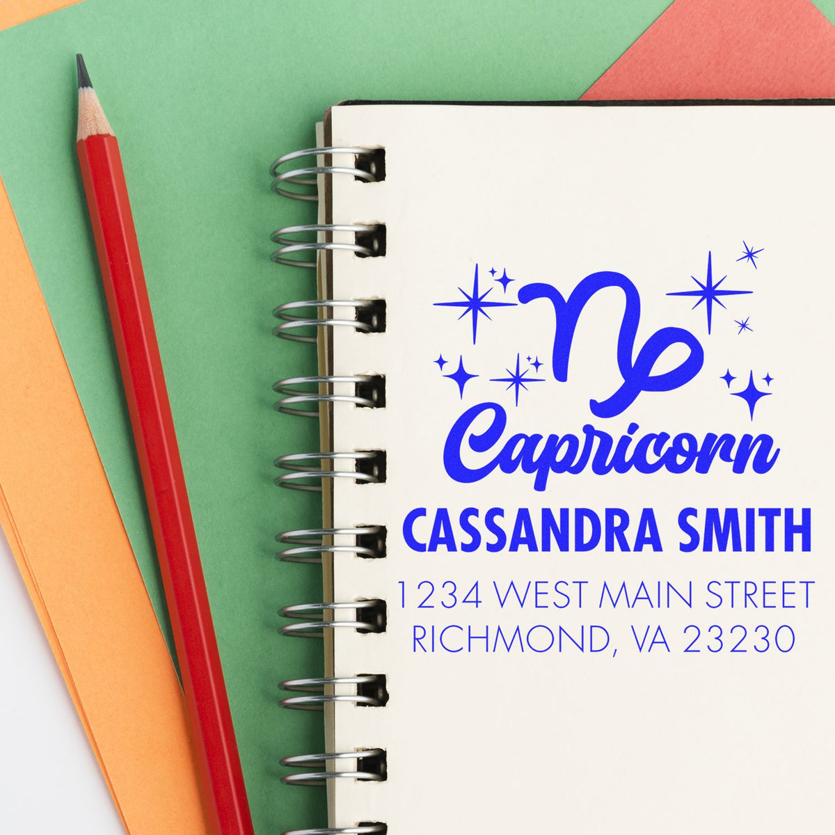 Custom blue Capricorn stamp and seal on a spiral notebook, with sparkles, Cassandra Smith name, address, and a red pencil on green paper.