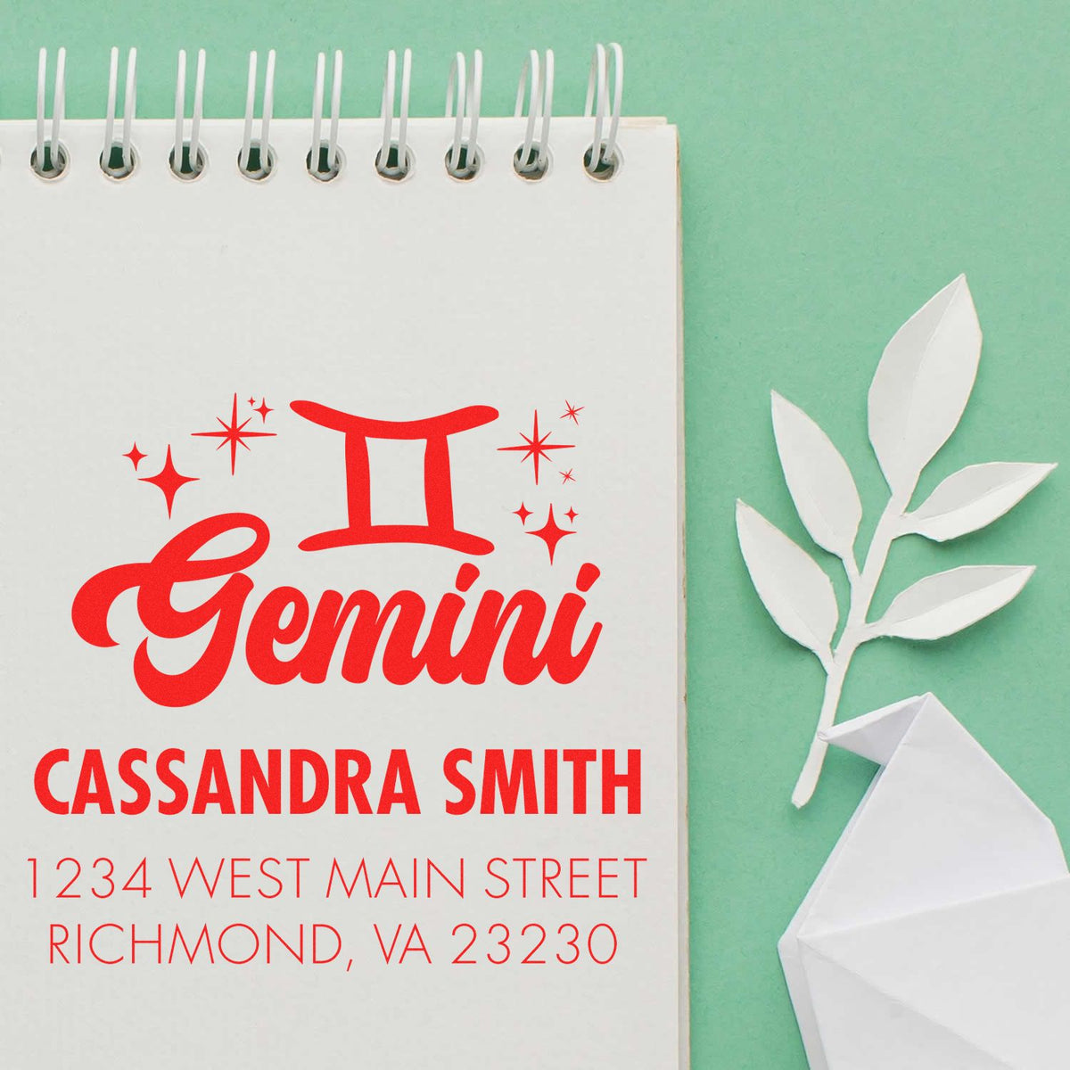 Custom red Gemini stamp and seal with Cassandra Smith address on white spiral notepad, mint green background.