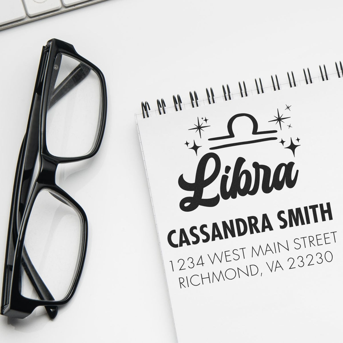 Black eyeglasses beside a custom Libra stamp and seal on a white spiral notepad with address.