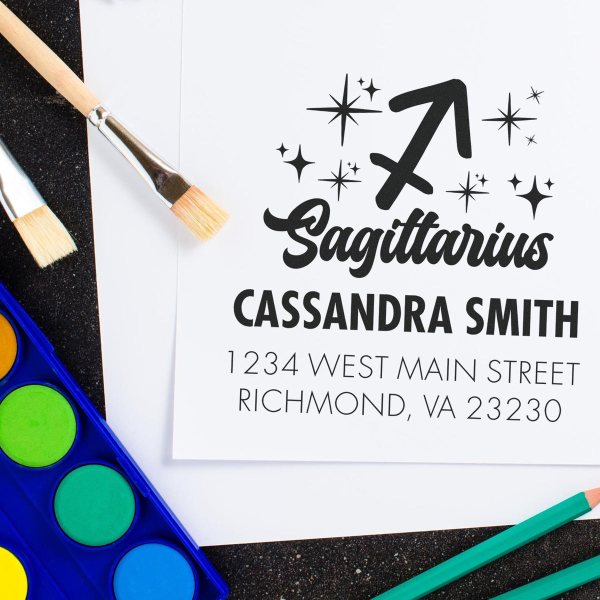 Black Sagittarius stamp seal with stars on white paper, beside paintbrushes and colorful art supplies.