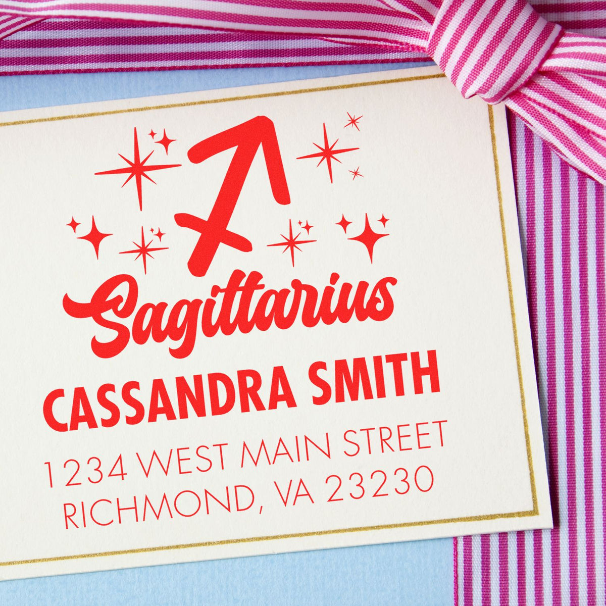 Red Sagittarius custom stamp seal with sparkles, name and address on cream card, framed by pink striped ribbon.