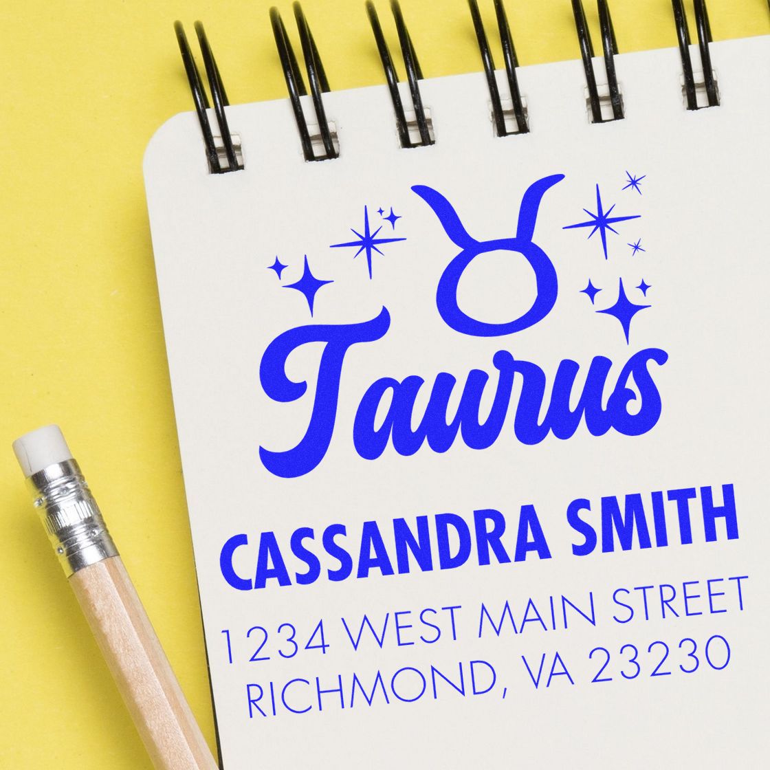 Blue Taurus custom stamp and address seal on white spiral notebook with yellow background and pencil.