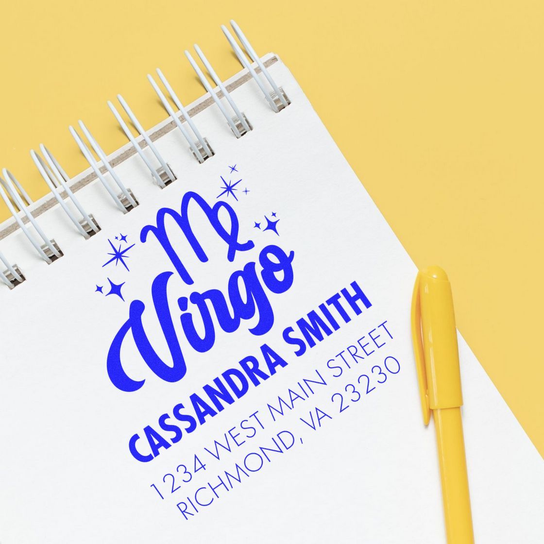 Blue custom stamp and seal design for Mr Virgo with address on a white spiral notebook, plus a yellow pen on a yellow background.