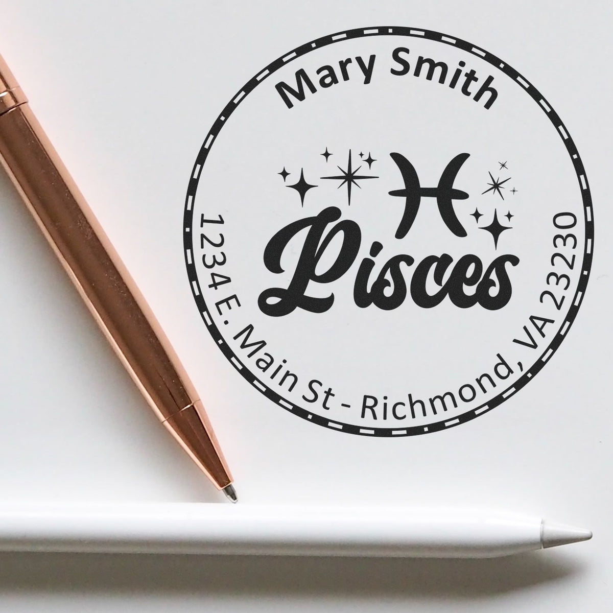 Black circular Pisces custom stamp seal reading Mary Smith, with rose-gold pen and white stylus on a white background.