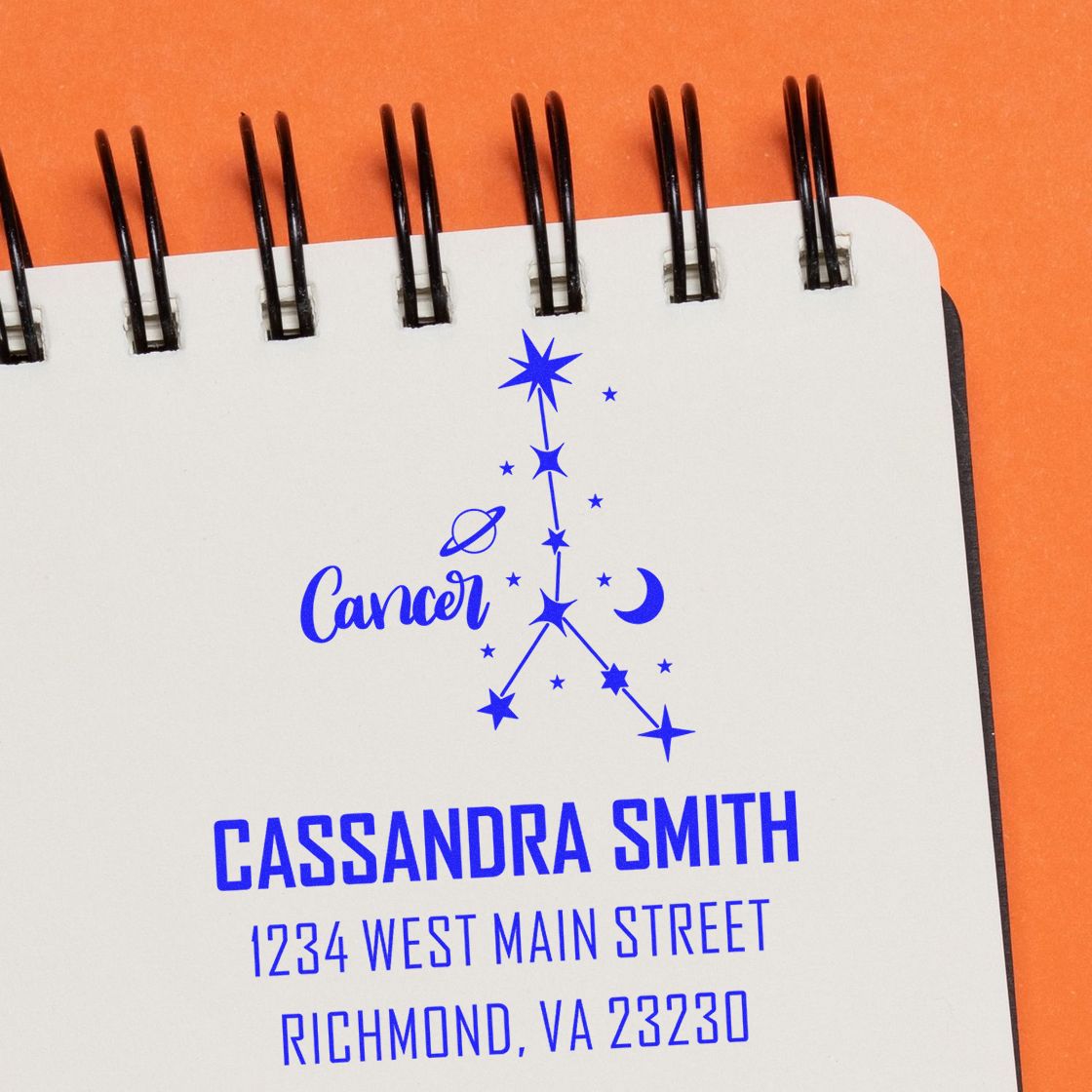Blue custom address stamp and Cancer zodiac seal on a white spiral notebook with black binding against an orange background.
