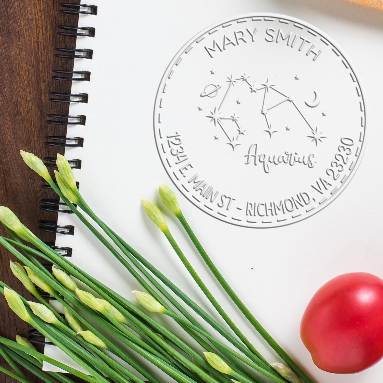 Embossed silver custom seal stamp on white notebook, with green stems and a red egg on a wood surface.