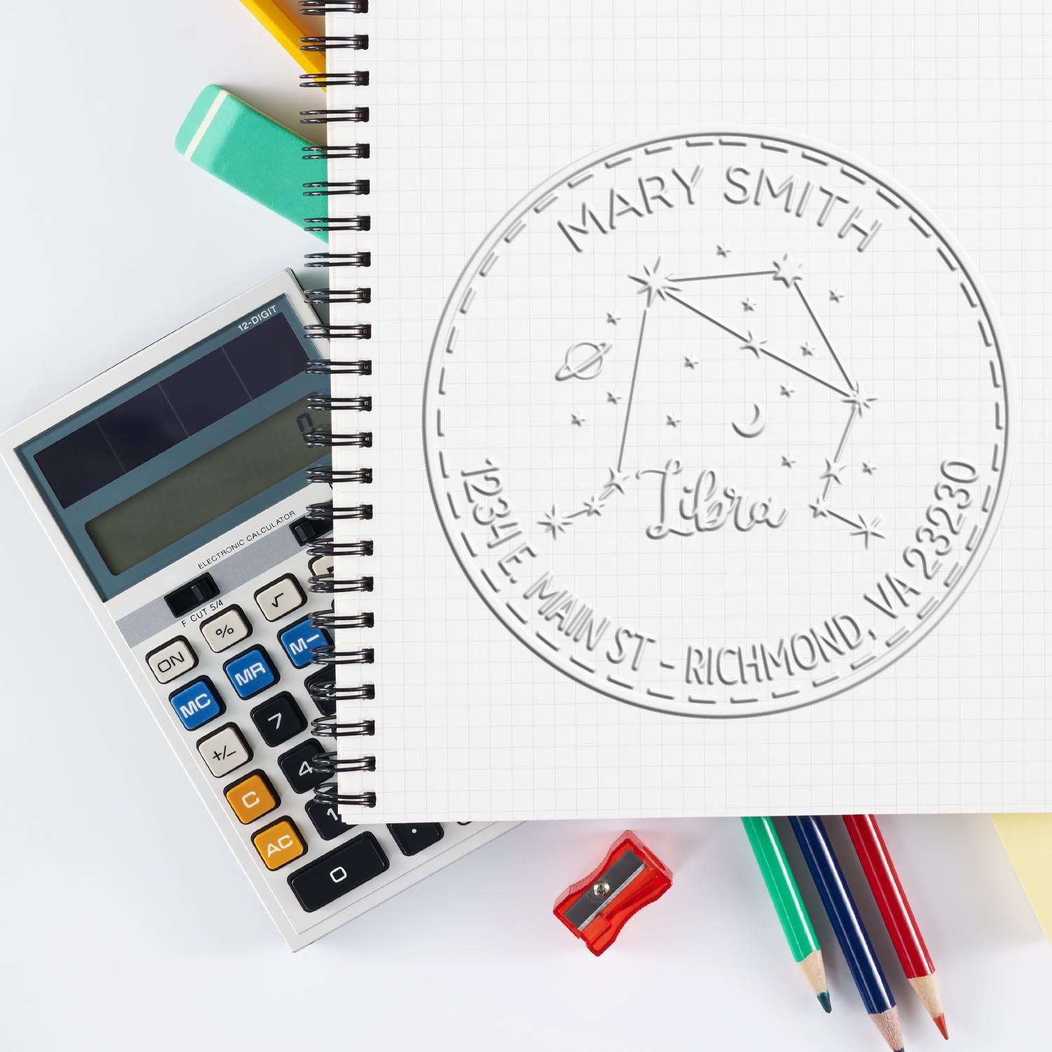 Silver embossed Libra seal stamp for Mary Smith on grid notebook, beside calculator, pencils, and sharpener.