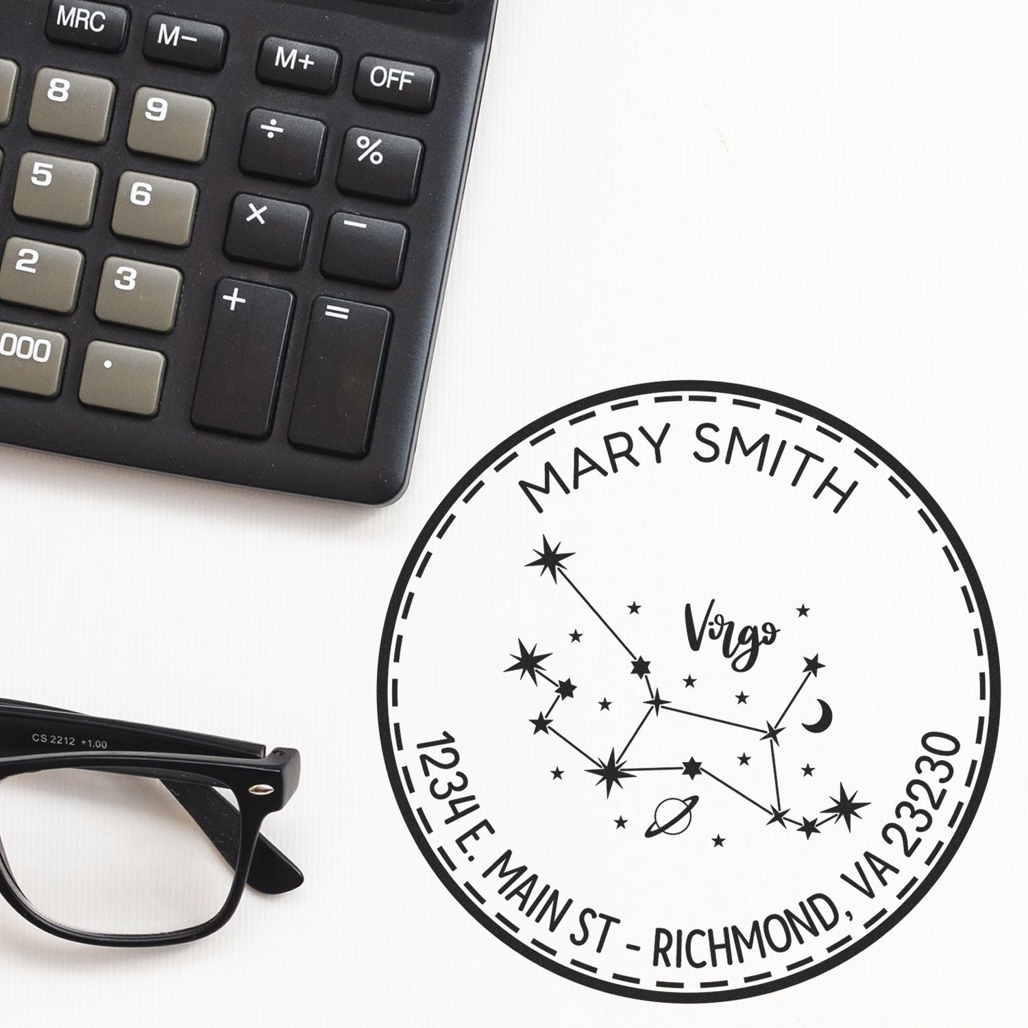 Black custom Virgo stamp and seal with constellation stars, on white background, beside a calculator and eyeglasses.