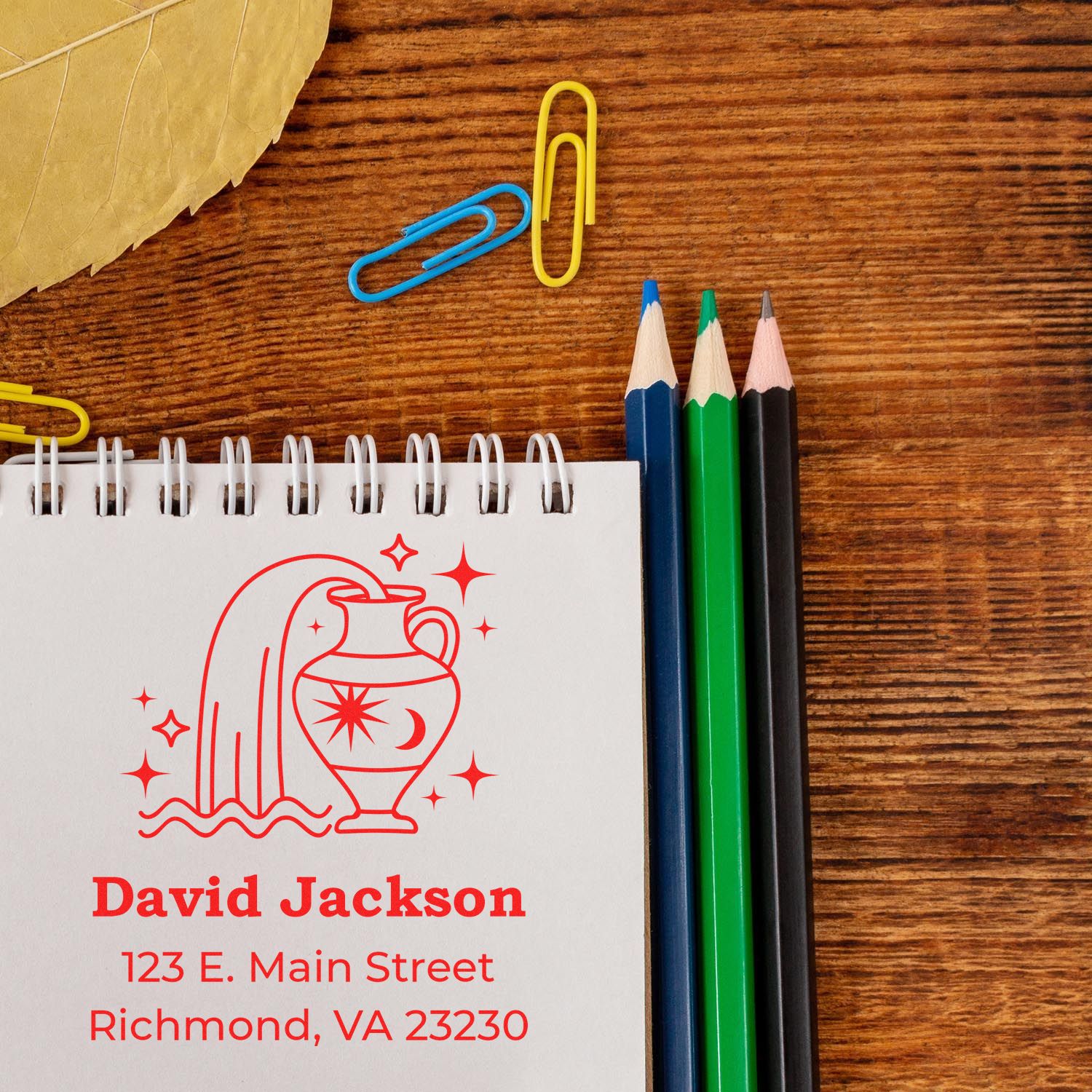 Custom red address stamp and seal with urn design on notebook, beside blue, green, and black pencils on a wood desk.