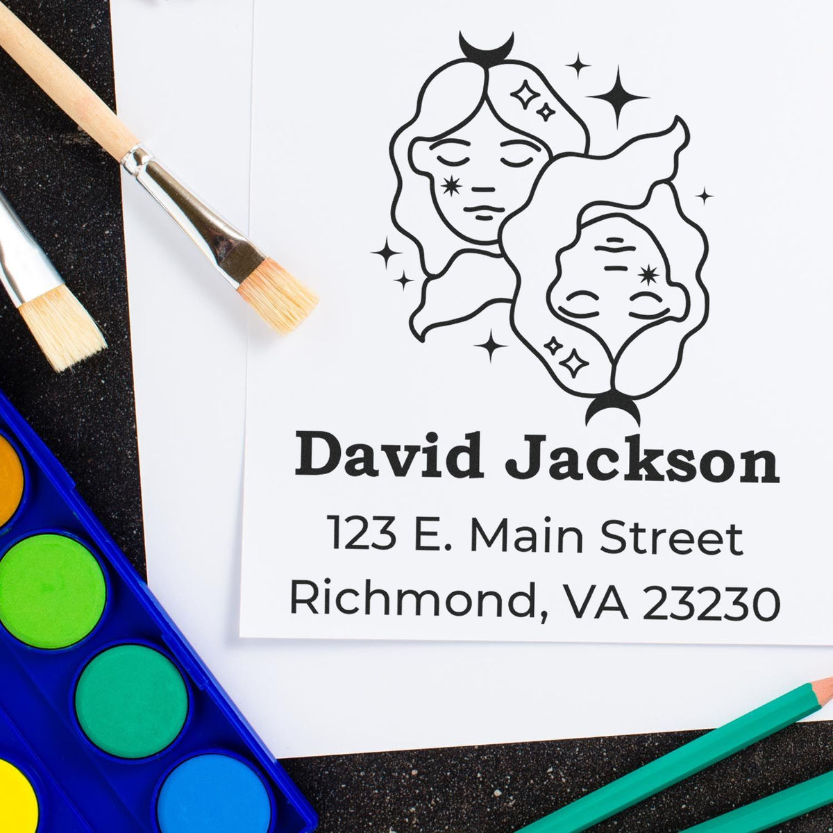Personalized black stamp and seal design with celestial faces on white paper, surrounded by paintbrushes, blue palette, and teal pencils.