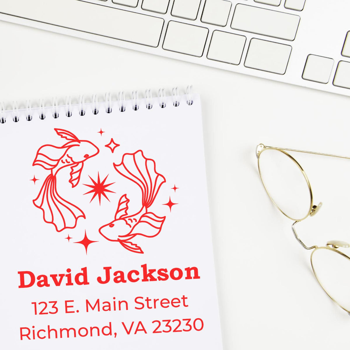 Red custom address stamp and seal design with goldfish artwork on white notebook, beside gold eyeglasses and keyboard