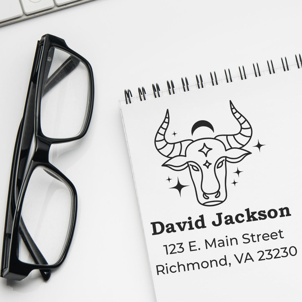 Black glasses beside a custom stamp and seal notebook with a bull head design, David Jackson name, and Richmond address on white background.