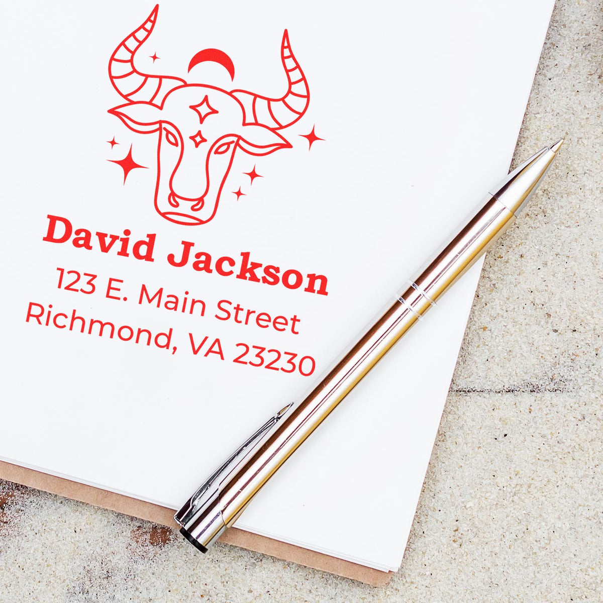 Custom red bull-head stamp and seal in red with name and address on white paper, beside a silver pen.