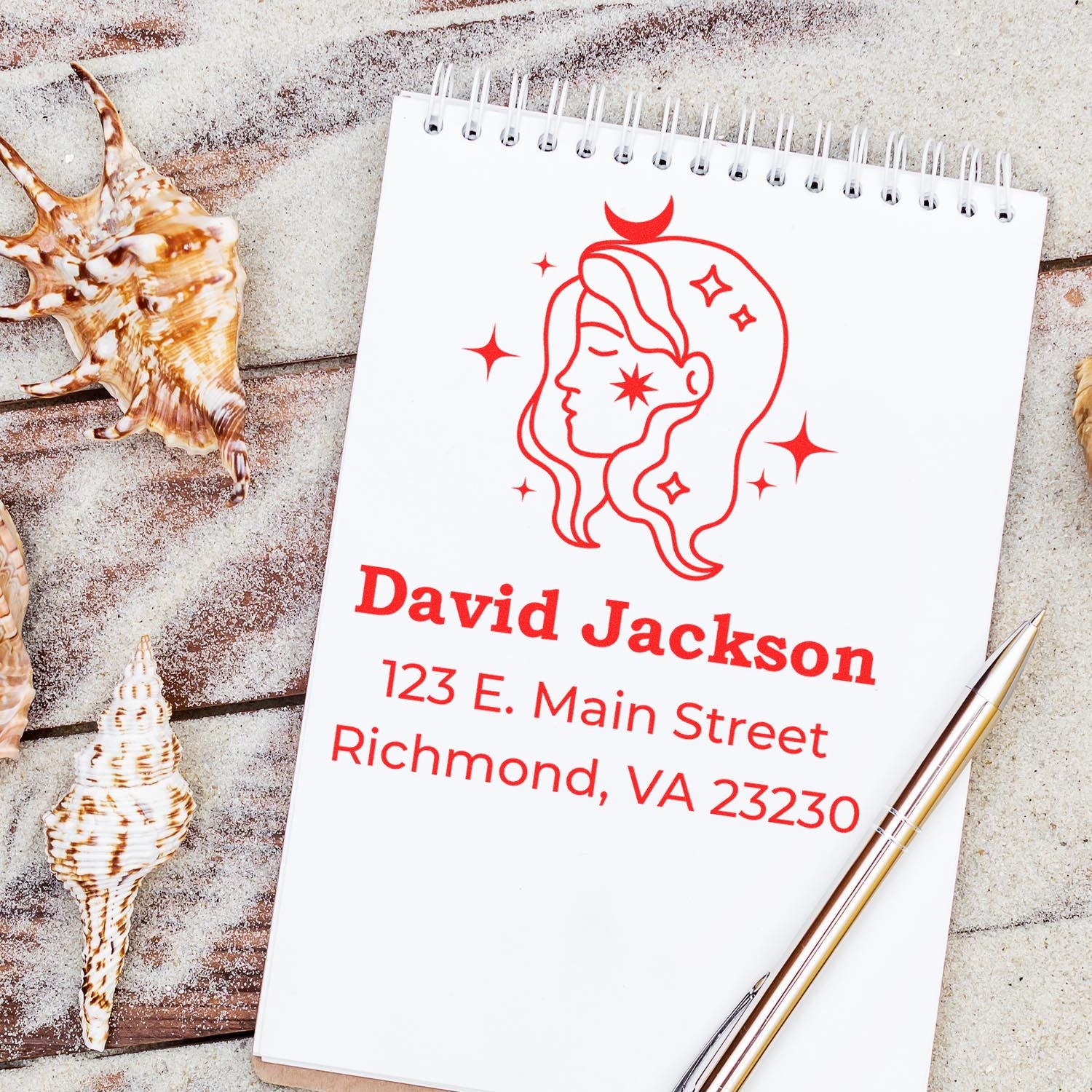 Custom red stamp and seal on white spiral notepad with name and address, beside seashells and a gold pen.