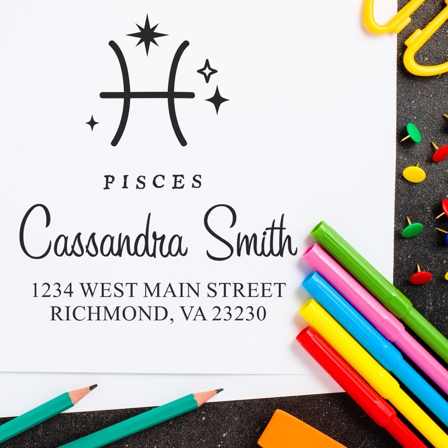 Pisces stamp and seal with black custom name and address, surrounded by colorful pens, pencils, and push pins.