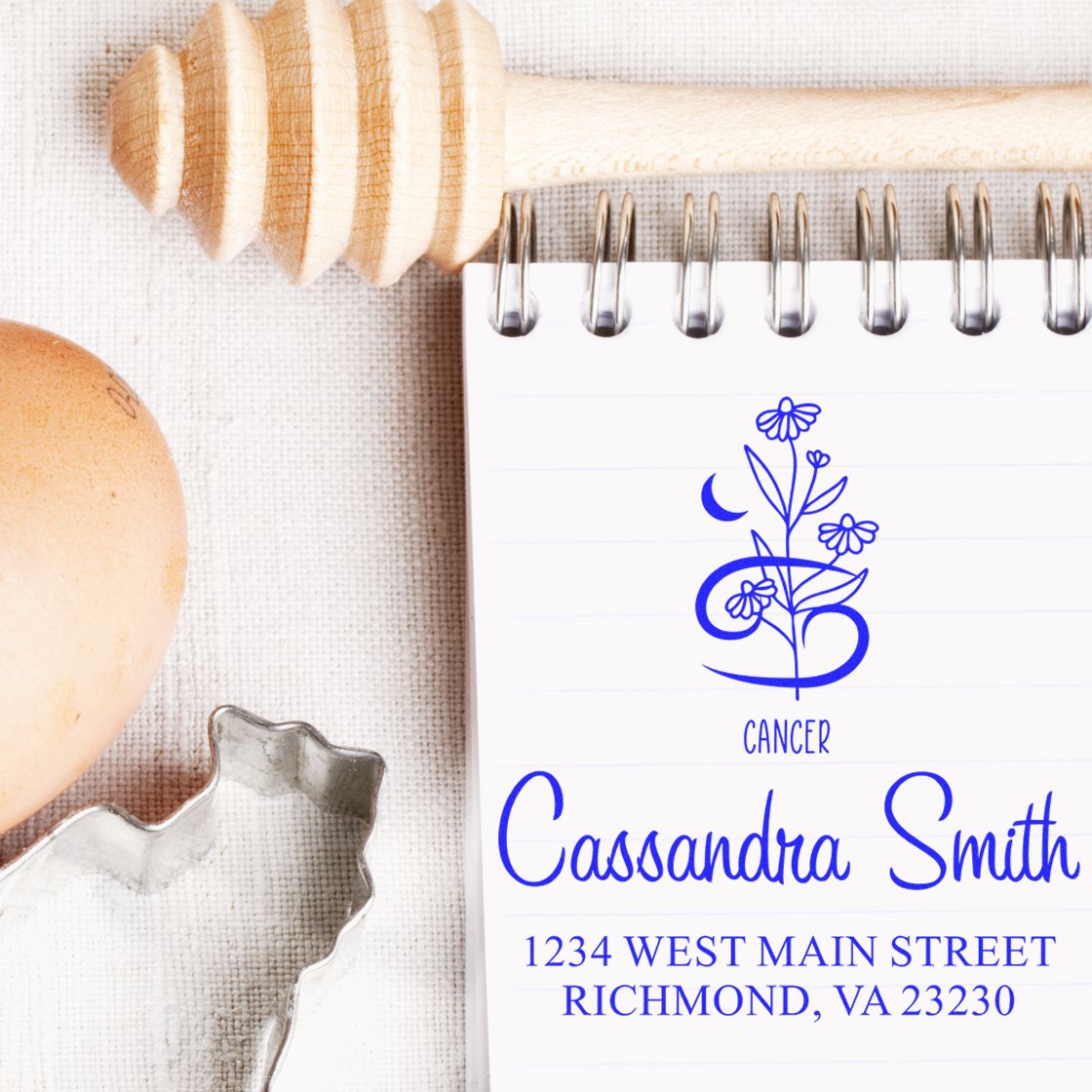 Blue custom stamp and seal products with Cancer floral design on a spiral notebook, beside a wooden honey dipper, egg, and metal cutter.