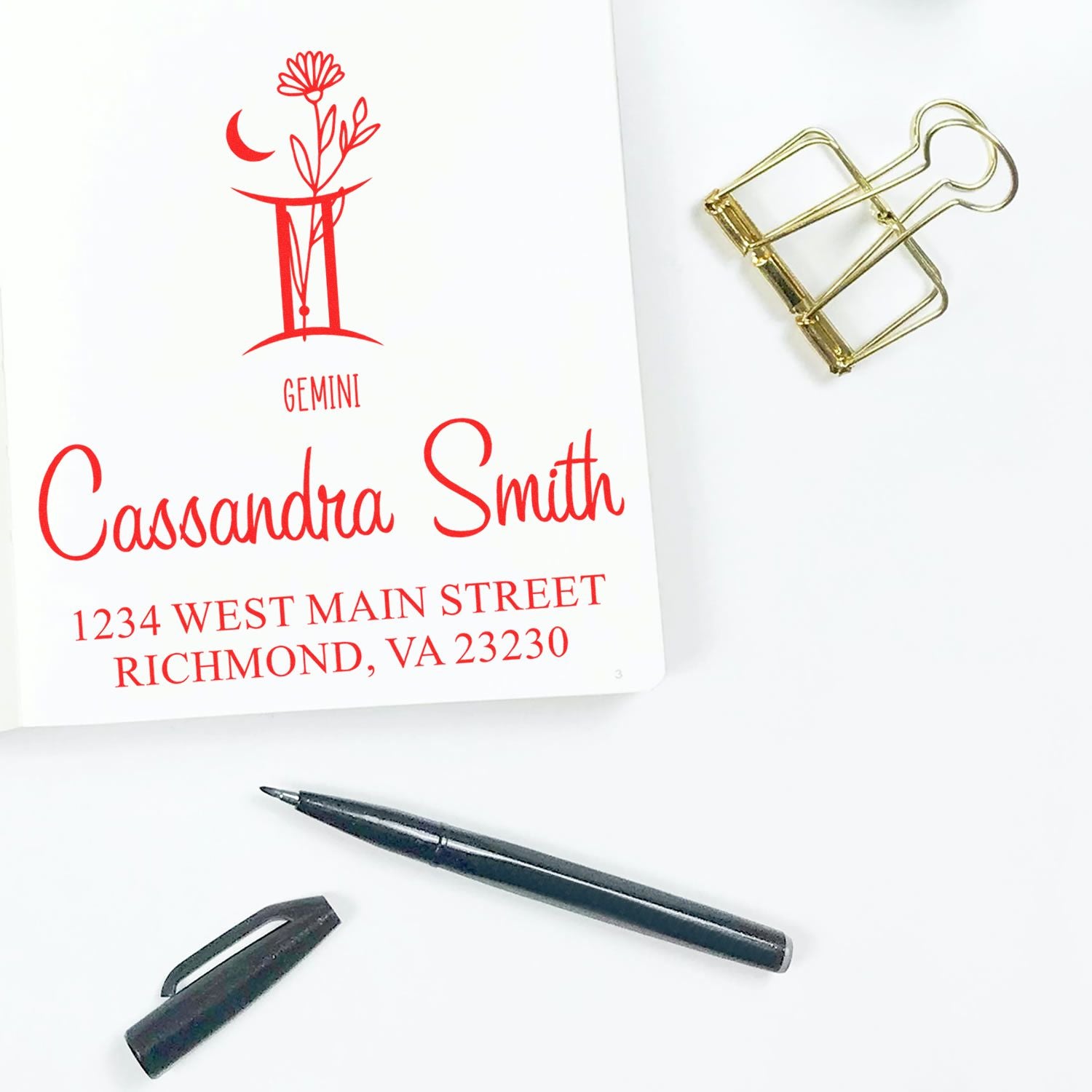 Red custom stamp and seal proof for Cassandra Smith, with Gemini design, on white paper; gold binder clip and black pen.