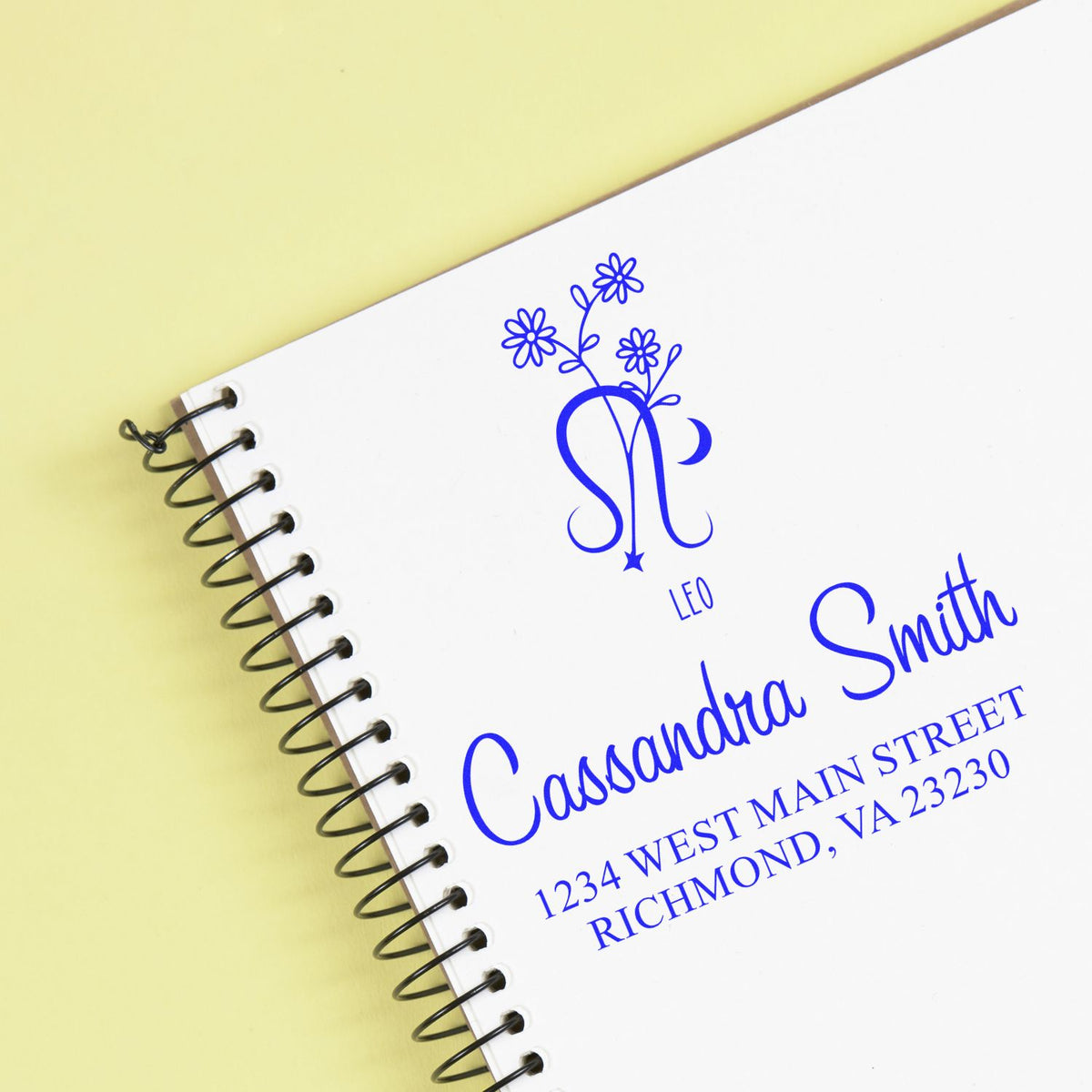 Blue personalized stamp and seal with floral monogram, name and address on a white spiral notebook against pale yellow.