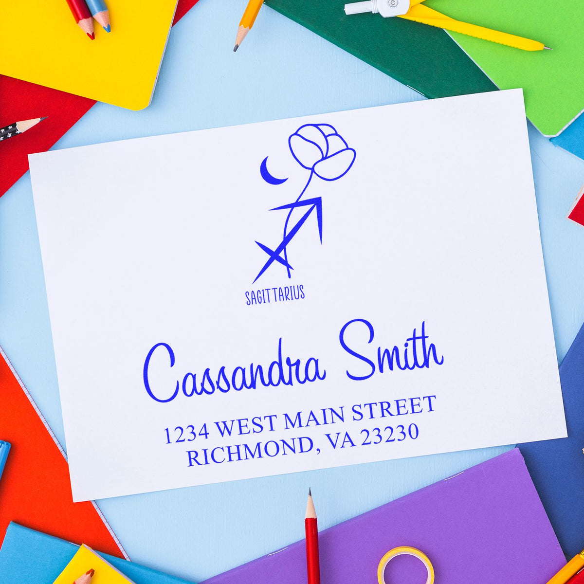 Blue Sagittarius return address stamp and seal on white card, surrounded by colorful notebooks and pencils.