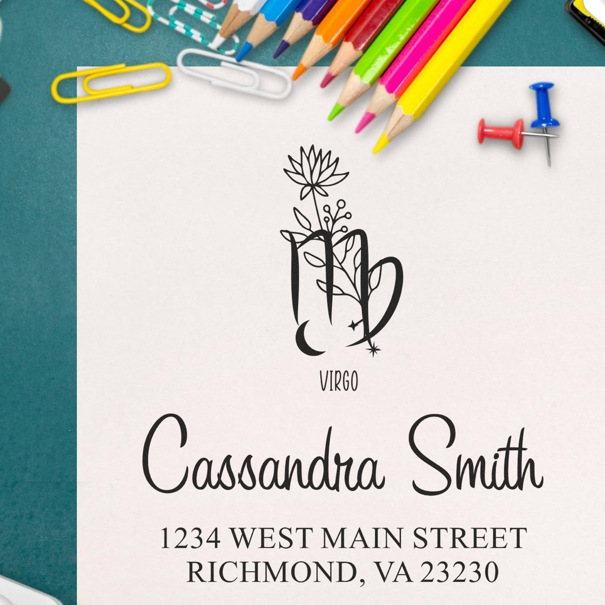 Custom stamp and seal design with Virgo floral monogram, black name and address, on white paper with colorful pencils.