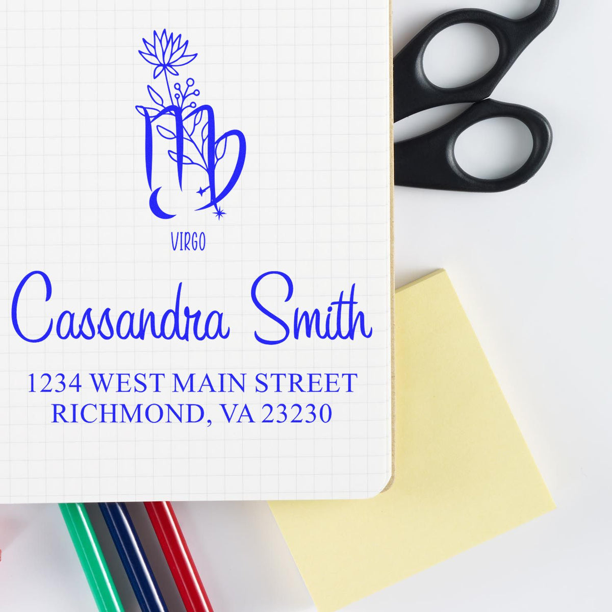 Blue Virgo personalized stamp seal with Cassandra Smith name and address, beside black scissors, sticky note, and colored pens.