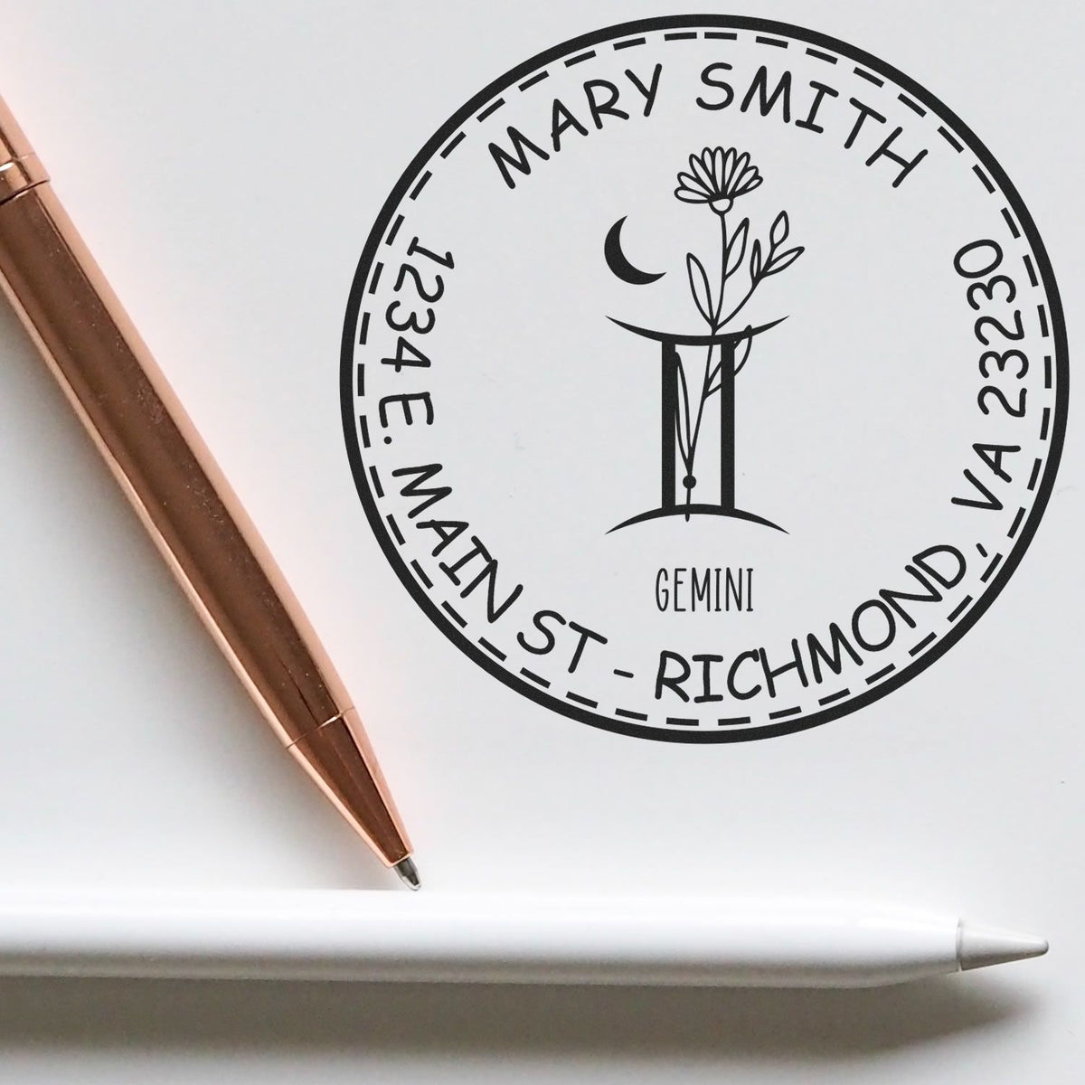Black circular custom stamp seal for Mary Smith, with Gemini floral emblem, on white paper beside a rose gold pen and white stylus.