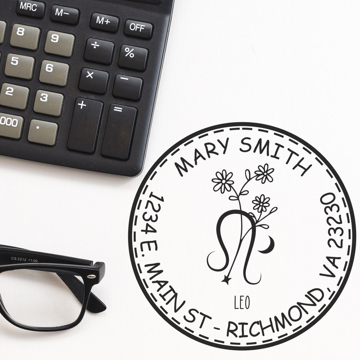 Circular black custom stamp and seal with floral monogram, Mary Smith, address details, beside calculator and glasses.