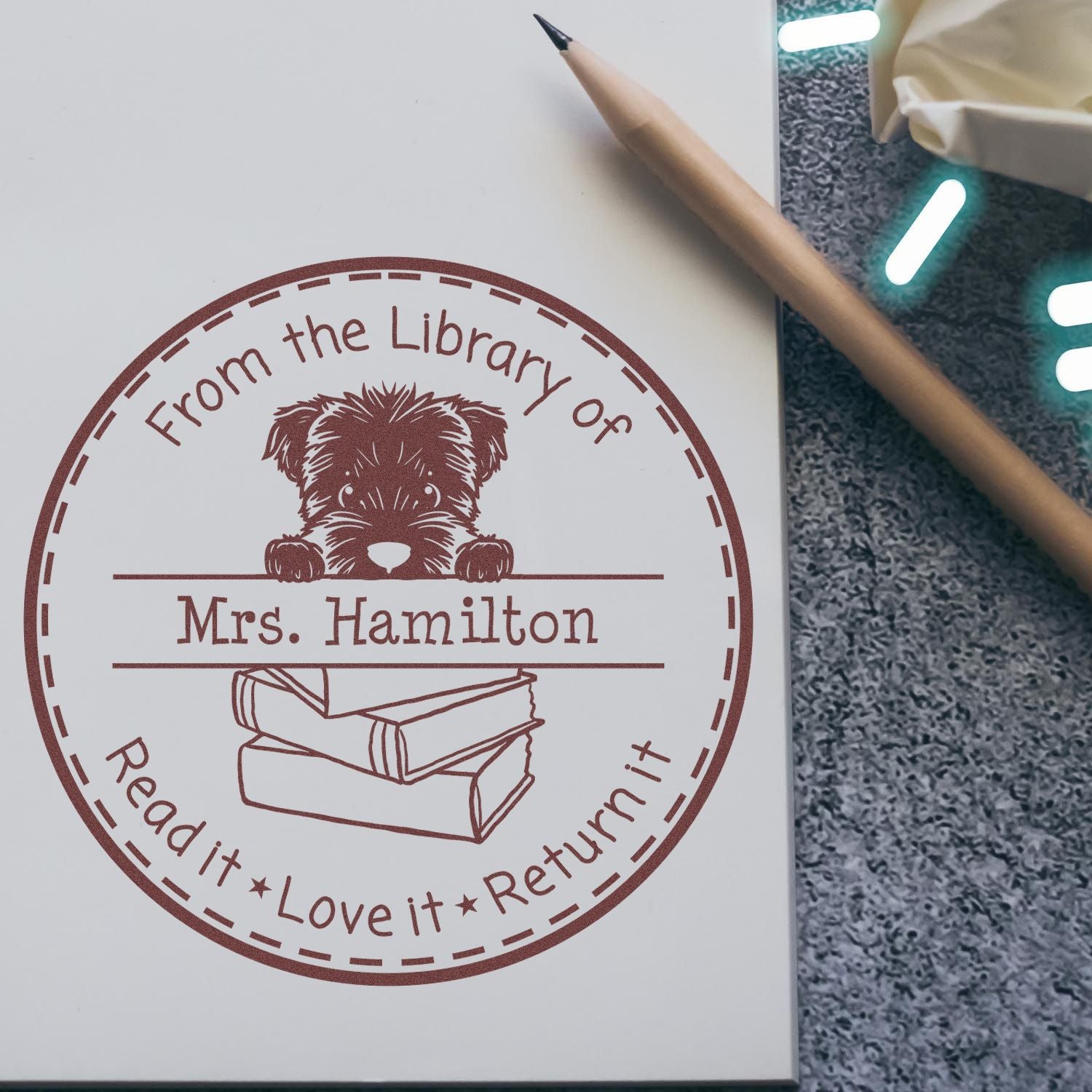 Brown custom library stamp seal with puppy over books, Mrs. Hamilton, on white paper beside a pencil.