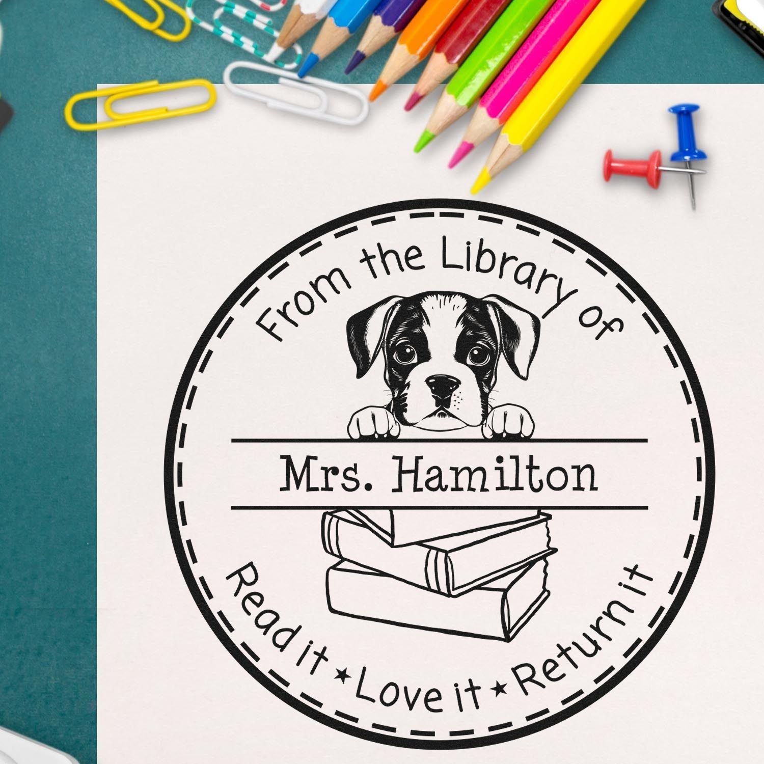 Black custom stamp and seal with puppy, books, and Mrs. Hamilton on white paper, framed by colorful pencils.
