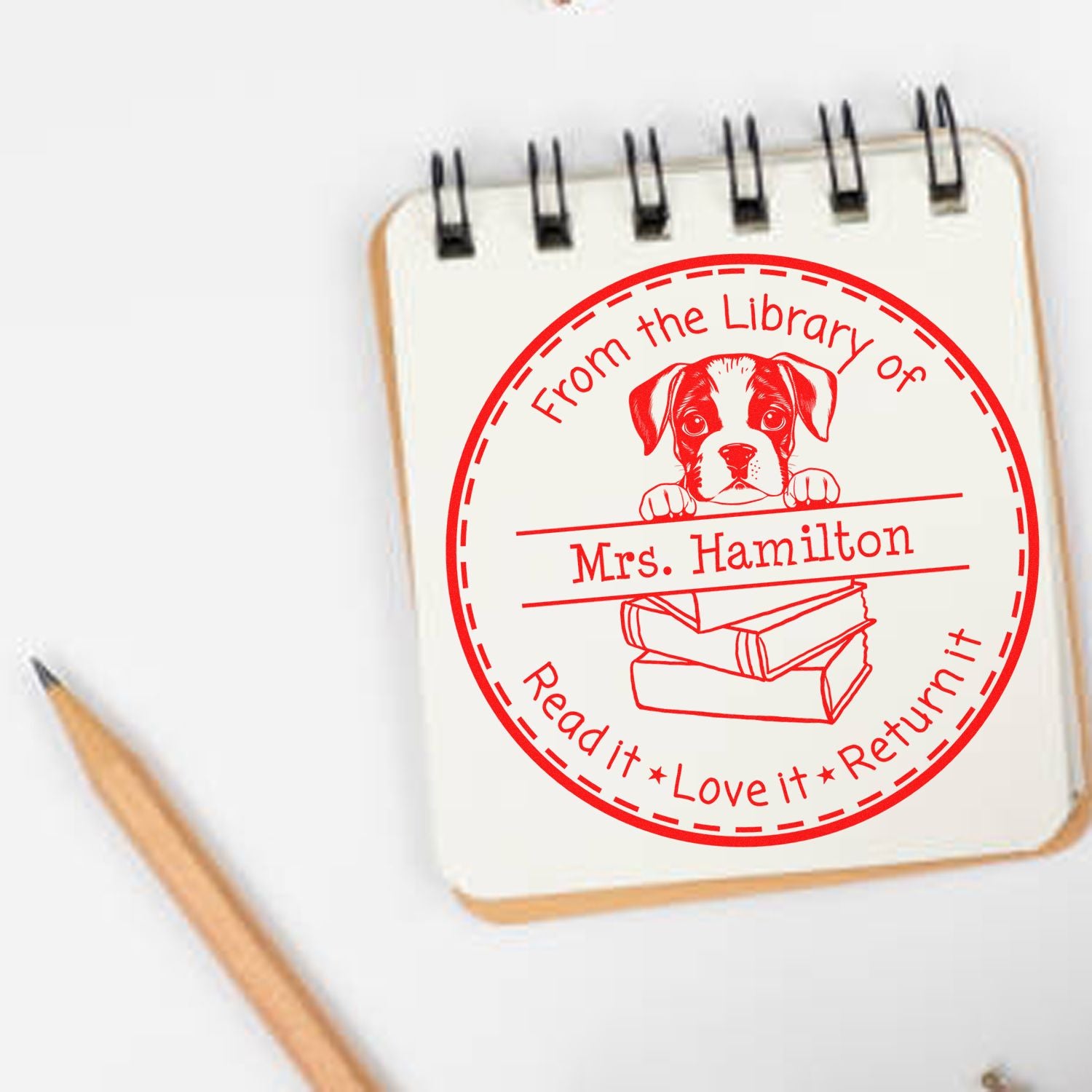 Red circular self-inking library return stamp with puppy and books, personalized Mrs. Hamilton, on a white spiral notebook.