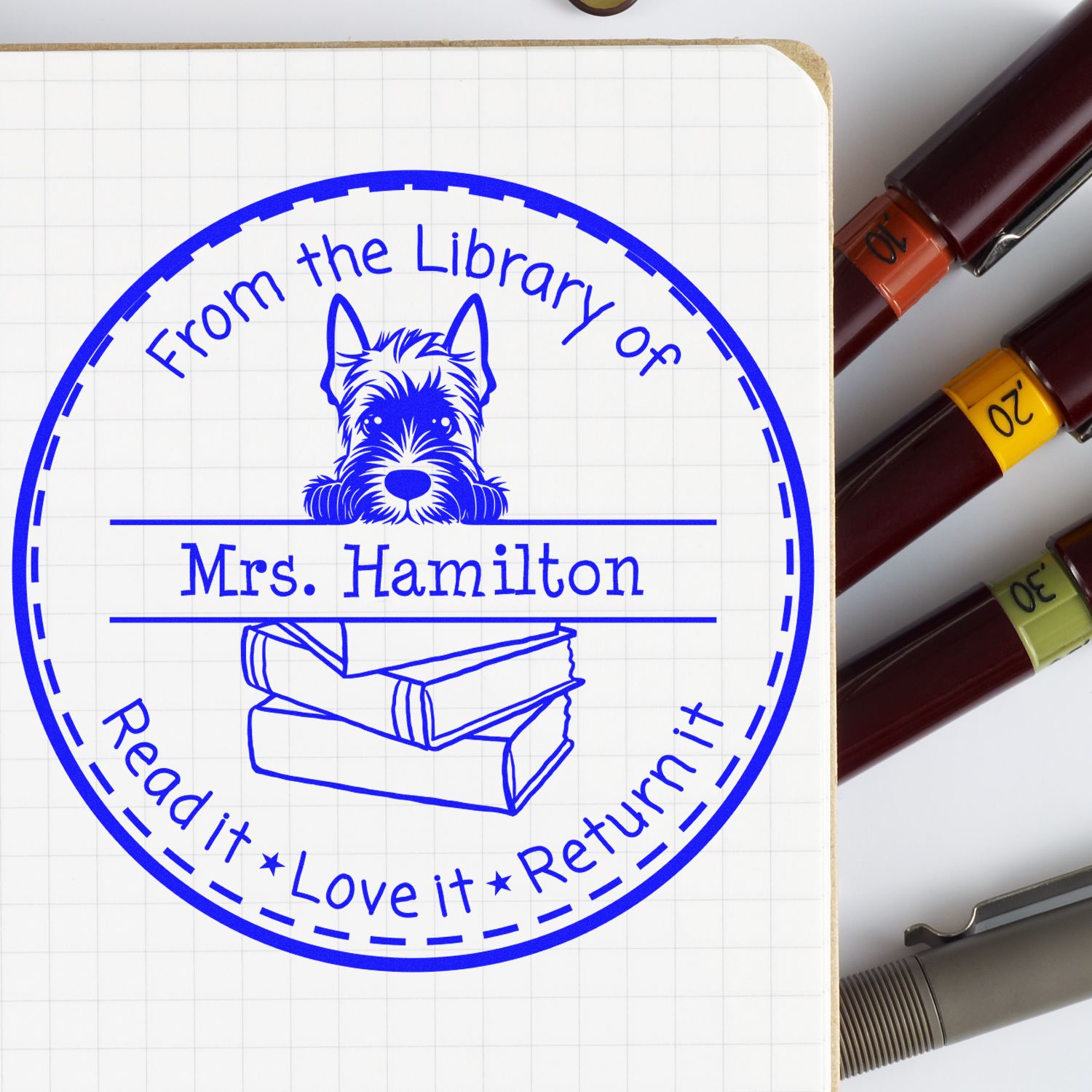 Blue custom library stamp seal with dog, books, and Mrs. Hamilton on grid paper beside colored markers.