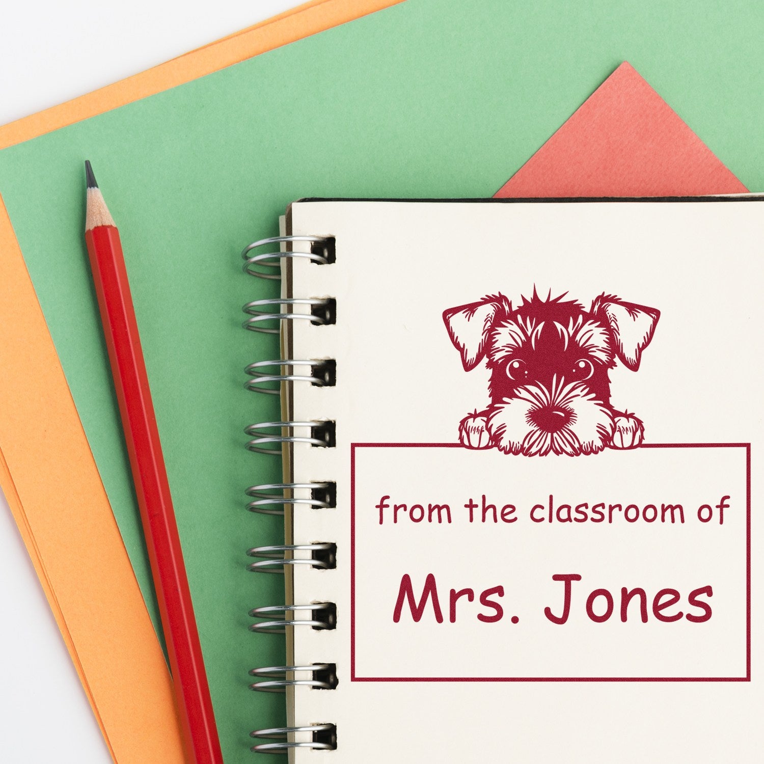 Custom red classroom stamp and seal with dog emblem and Mrs. Jones on spiral notebook, beside red pencil and green-orange paper