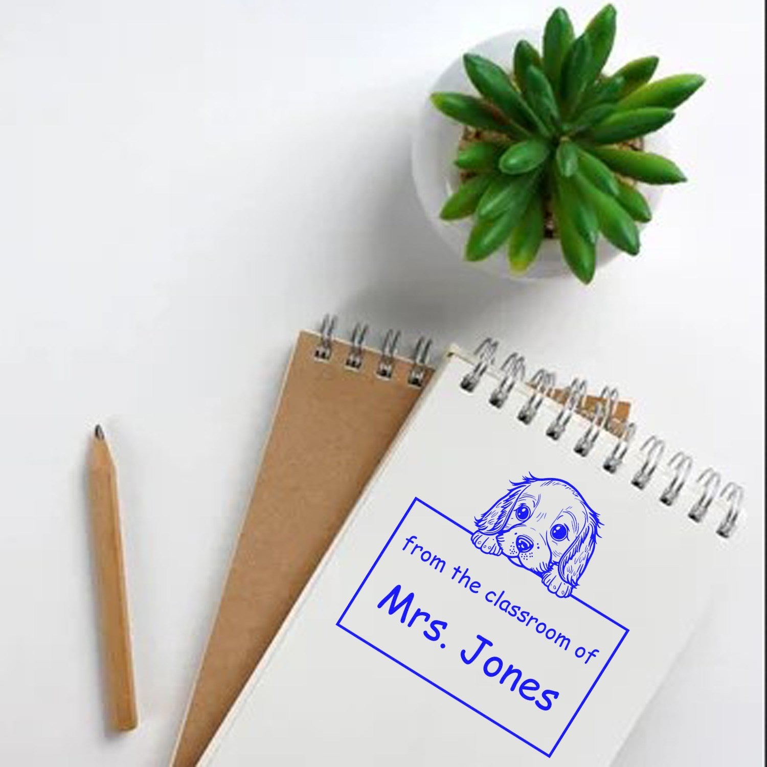 Blue custom stamp and seal classroom design on white spiral notebook, with dog illustration, Mrs. Jones text, pencil, and potted plant.