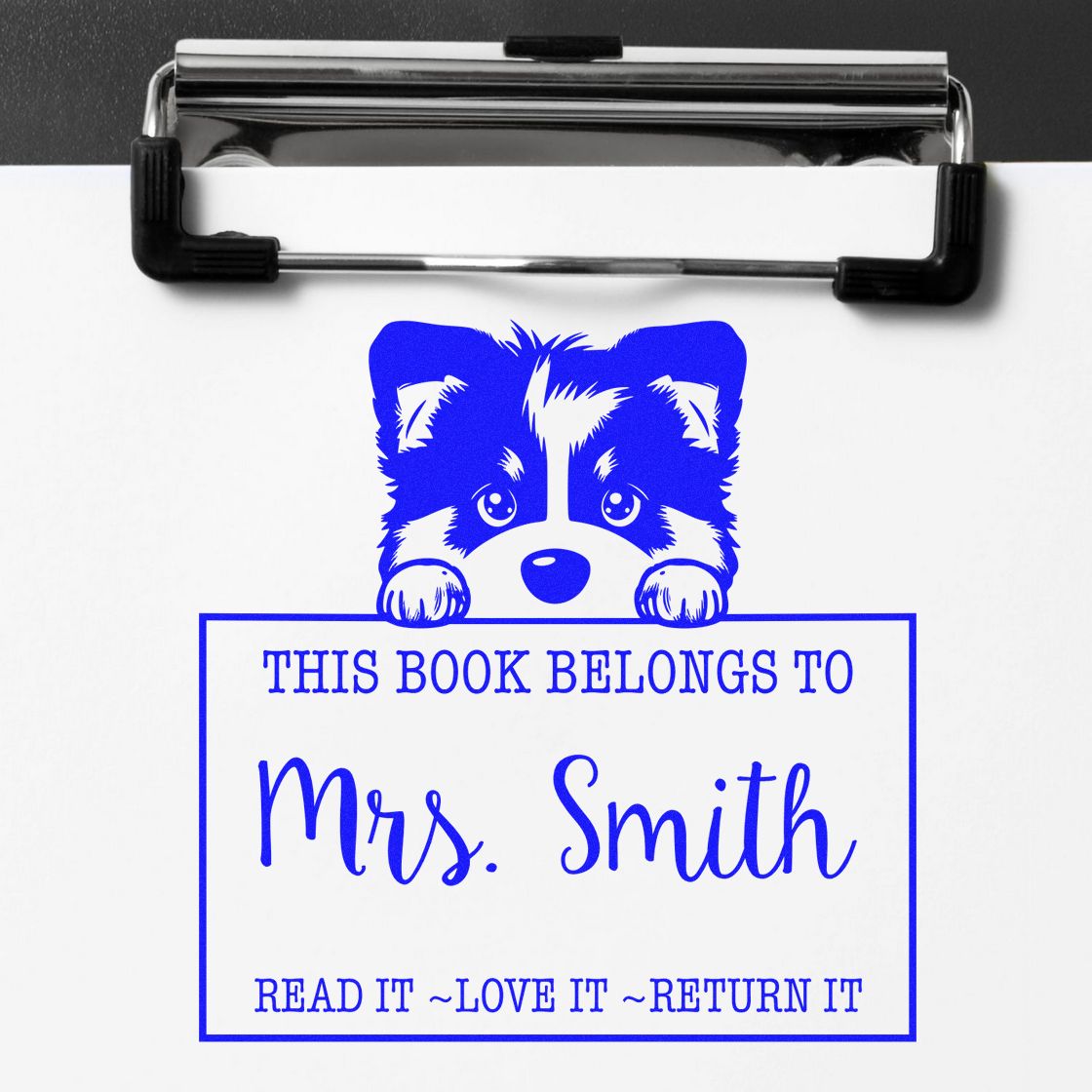 Blue custom book ownership stamp with puppy design and This Book Belongs to Mrs. Smith on white paper under a clipboard.