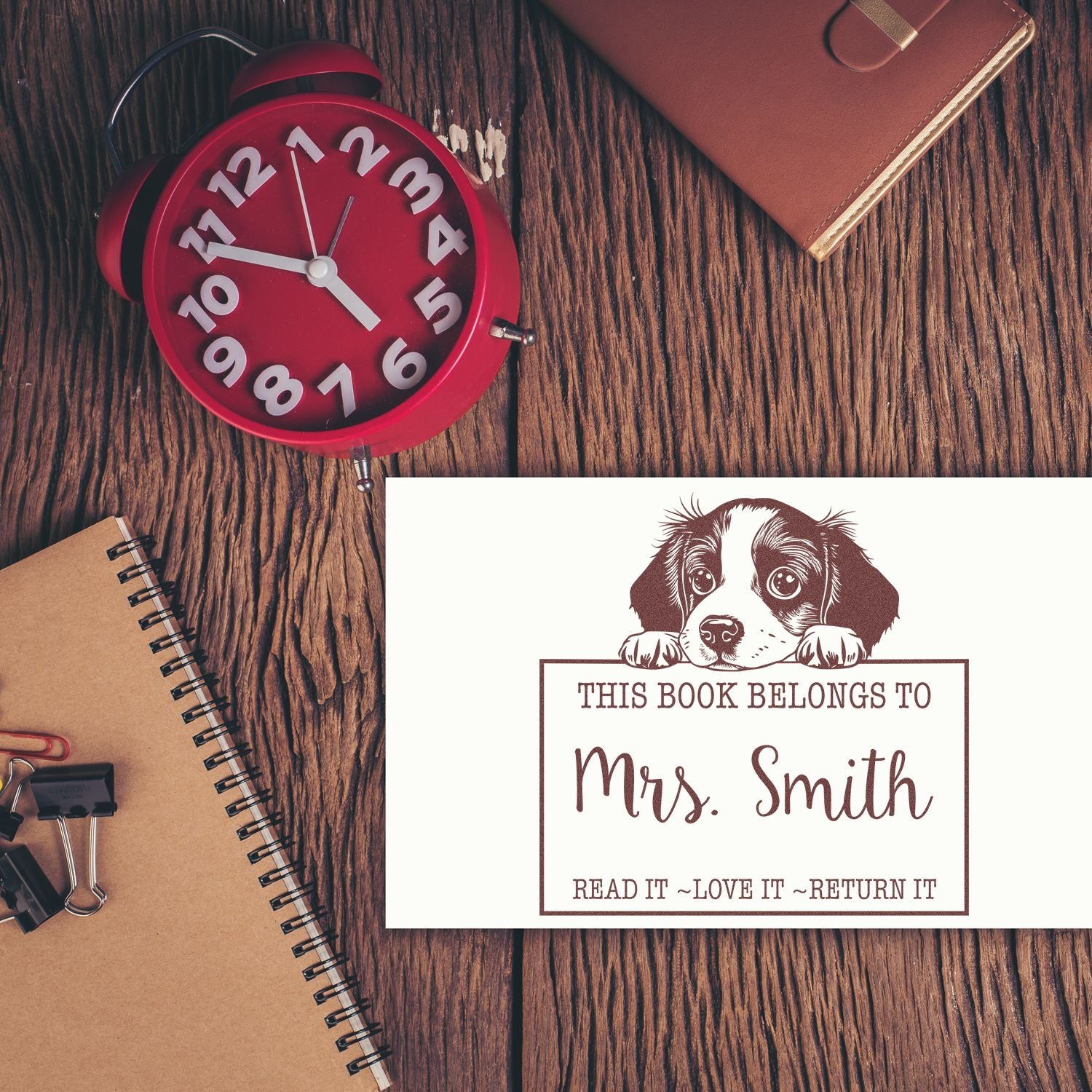 Brown custom book stamp and seal with dog, Mrs. Smith text on white card, beside a red alarm clock and notebook.