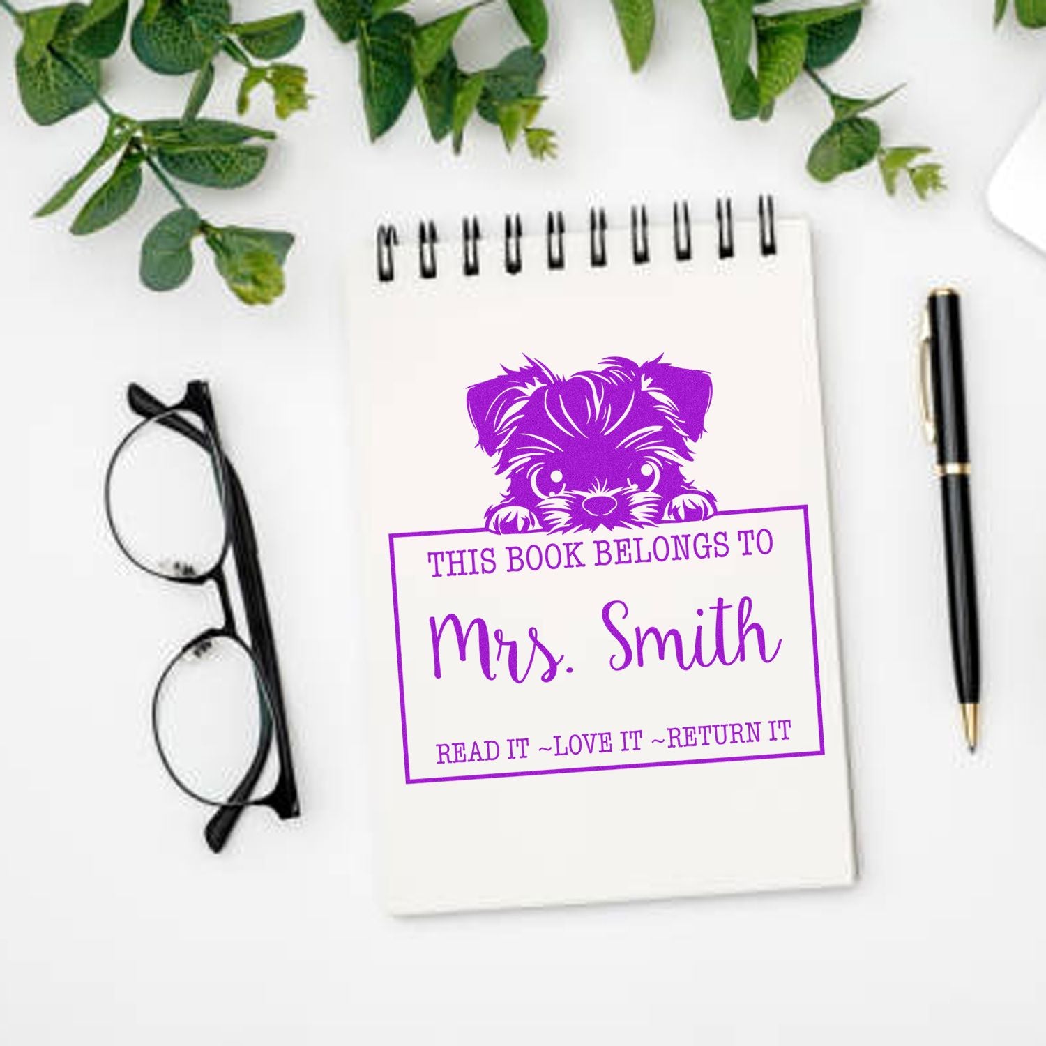 Purple custom book stamp and seal with puppy for Mrs. Smith on white notebook, with black glasses, pen, and green leaves.