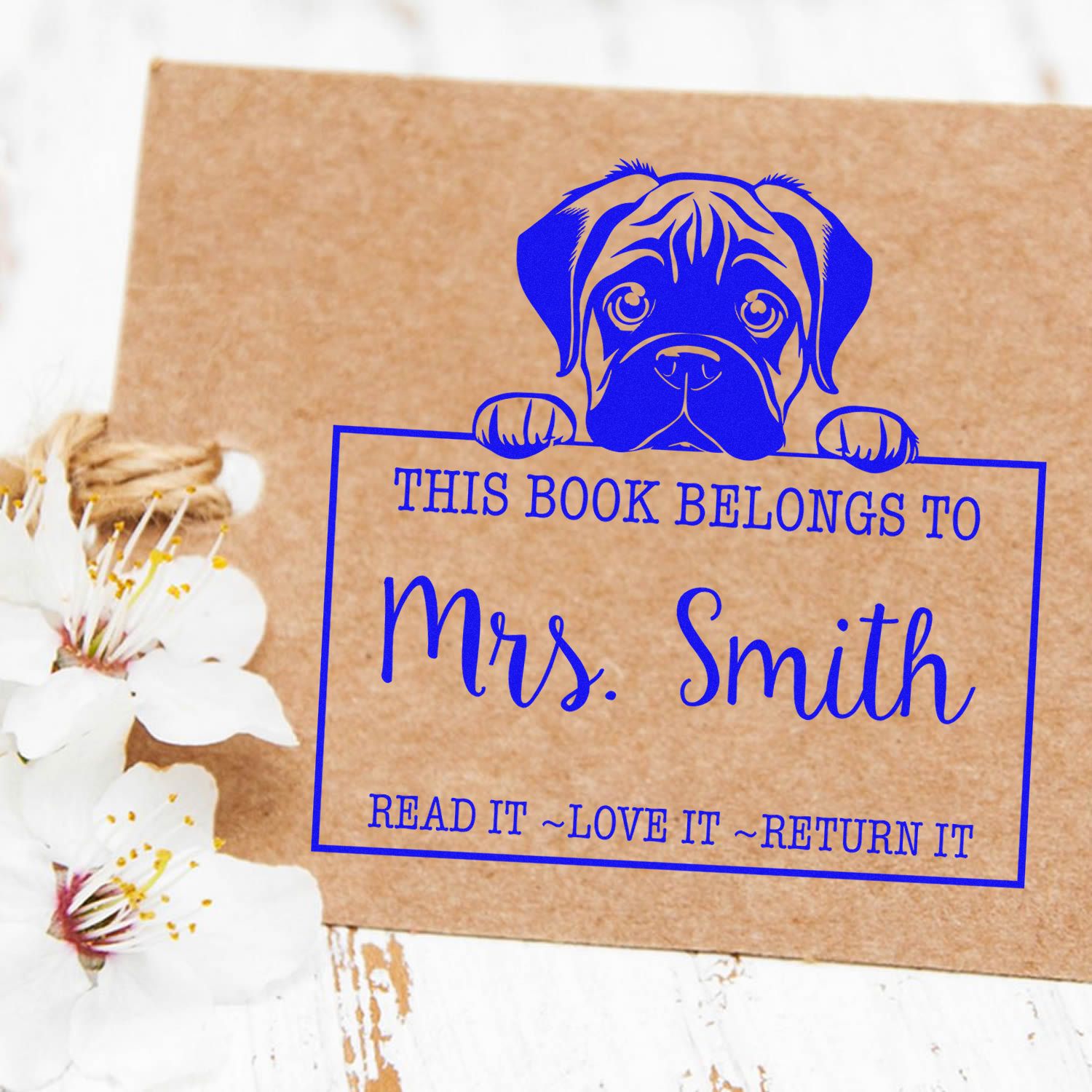 Blue personalized book stamp and seal with dog graphic, reading This book belongs to Mrs. Smith, on kraft paper with white flowers.