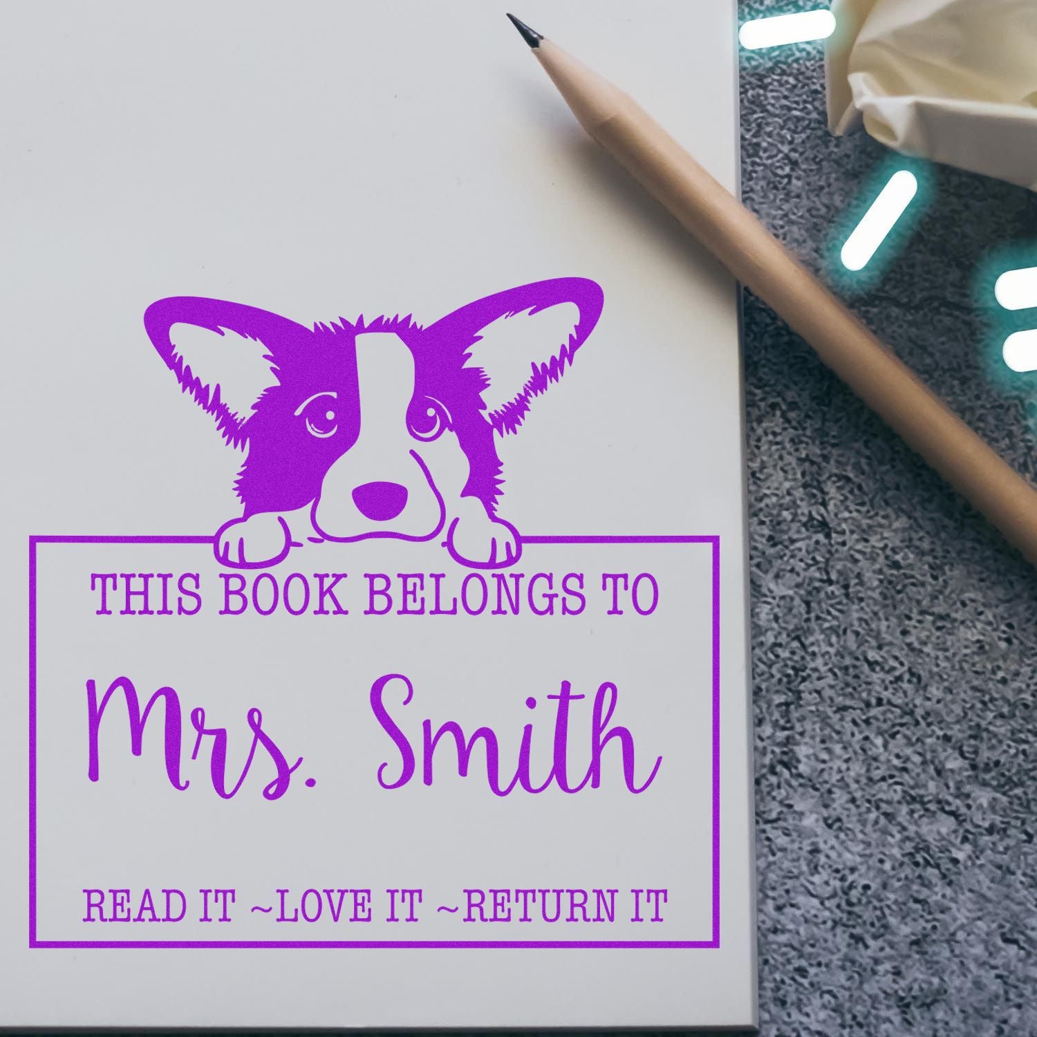 Purple corgi custom book stamp/seal reading This Book Belongs to Mrs. Smith on white paper, with a pencil nearby.