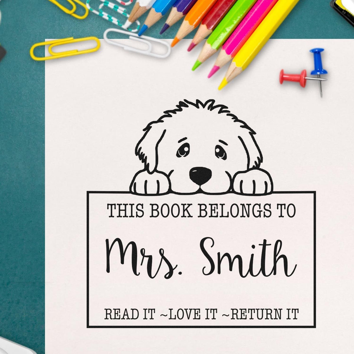 Personalized dog book stamp and seal in black, reading This Book Belongs to Mrs. Smith, with colorful pencils on white paper.