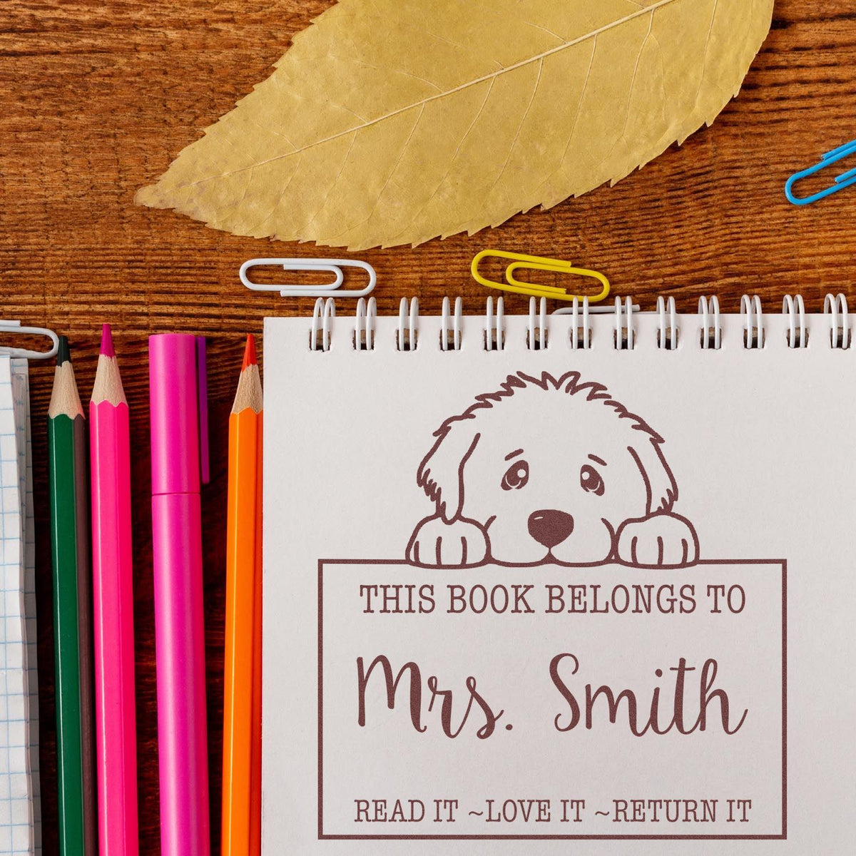 Personalized bookplate stamp seal with a brown dog design, Mrs. Smith text, and colorful pencils on a wood desk.