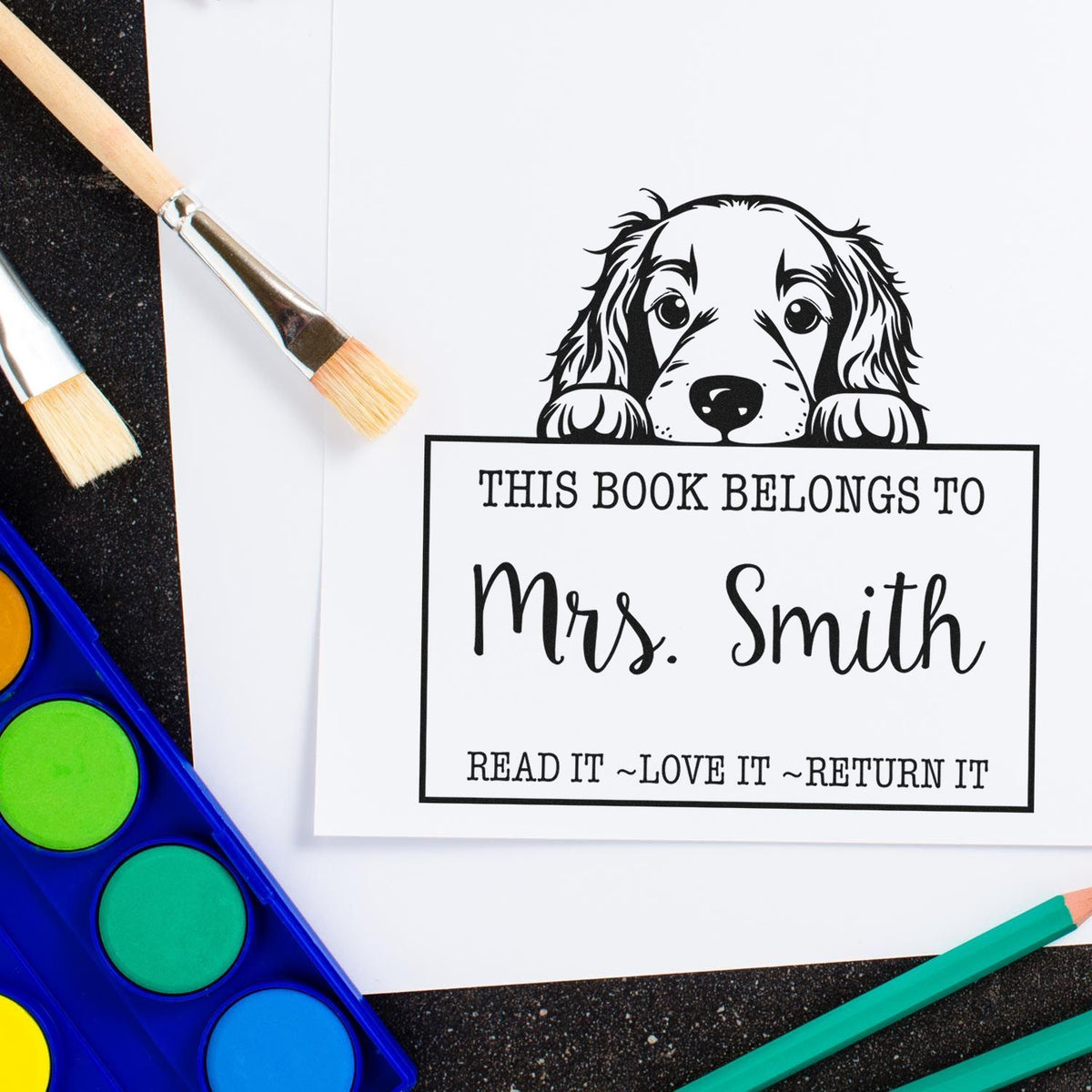 Black book stamp and seal for Mrs. Smith with dog illustration, on white paper beside paintbrushes and colorful paints.