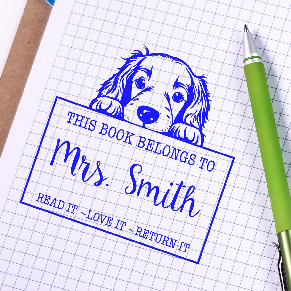 Blue custom stamp and seal with puppy graphic and This book belongs to Mrs. Smith on graph paper beside a green pen.