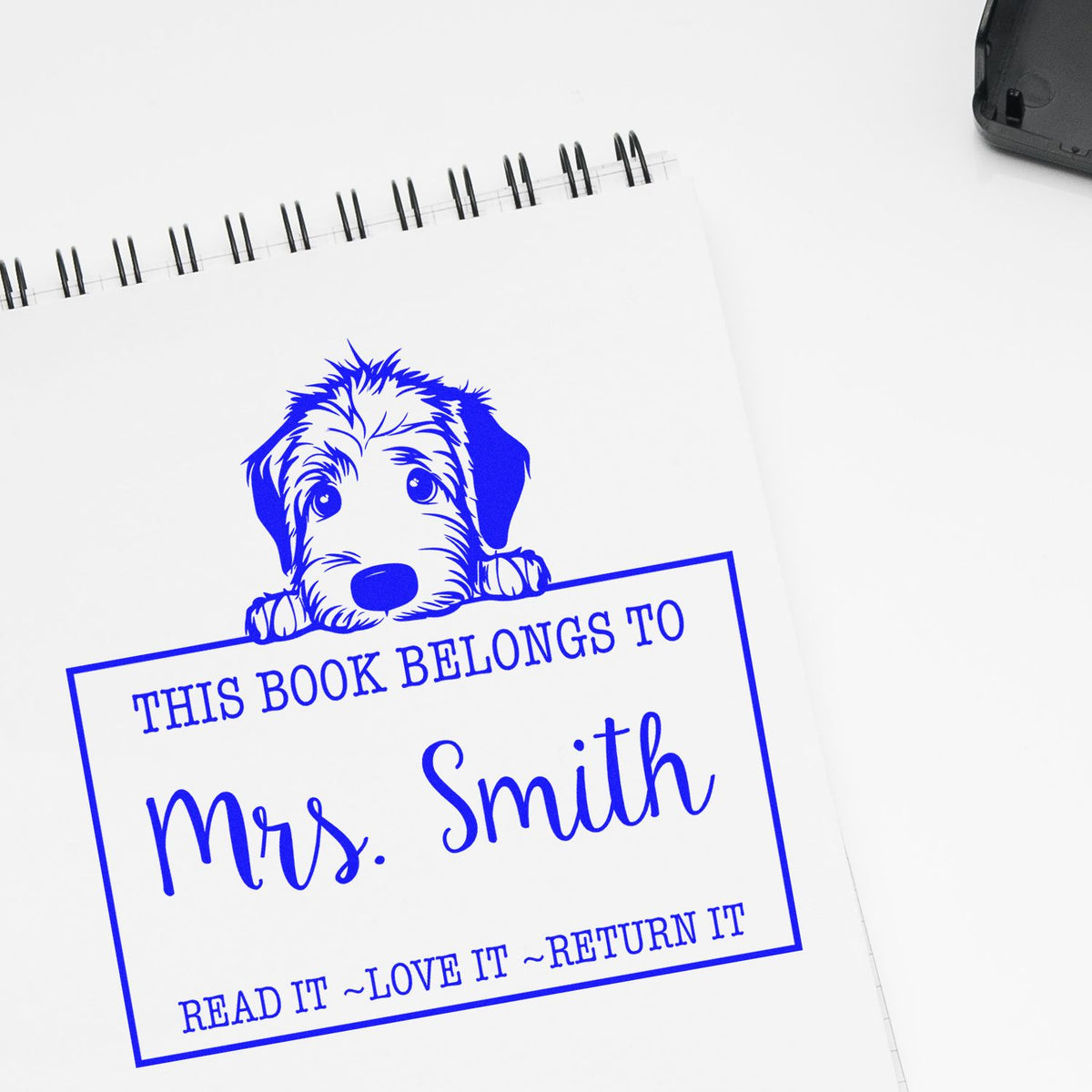 Blue custom book stamp and seal with dog graphic, reading This Book Belongs to Mrs. Smith, on a white notebook.