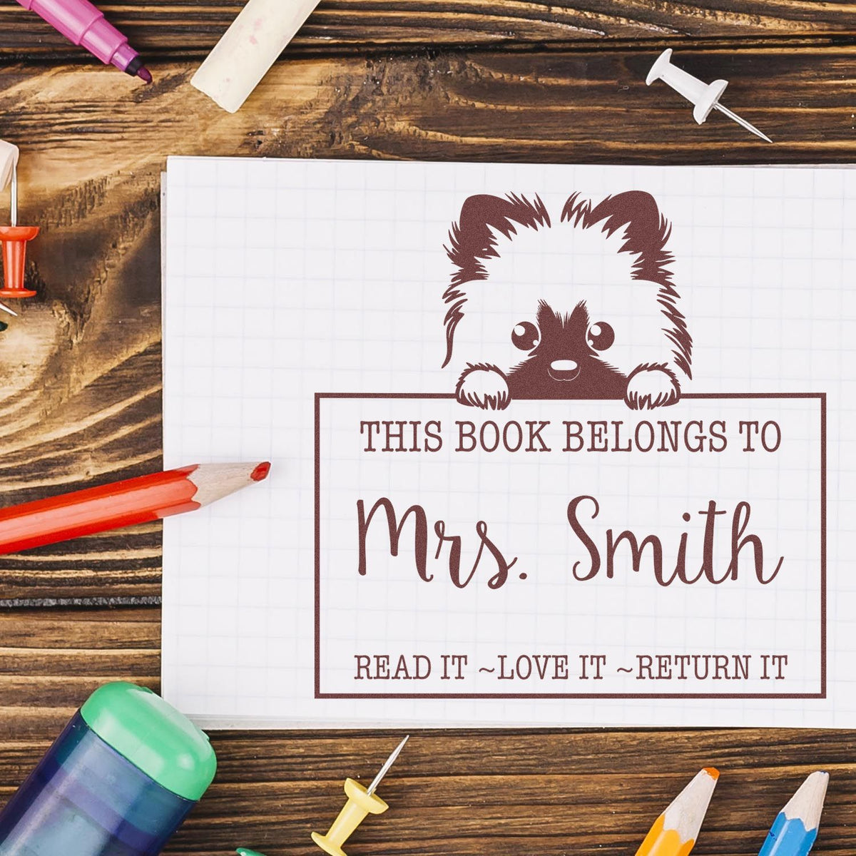 Brown bookplate stamp seal with bear, Mrs. Smith, on white notebook beside colored pencils on rustic wood.