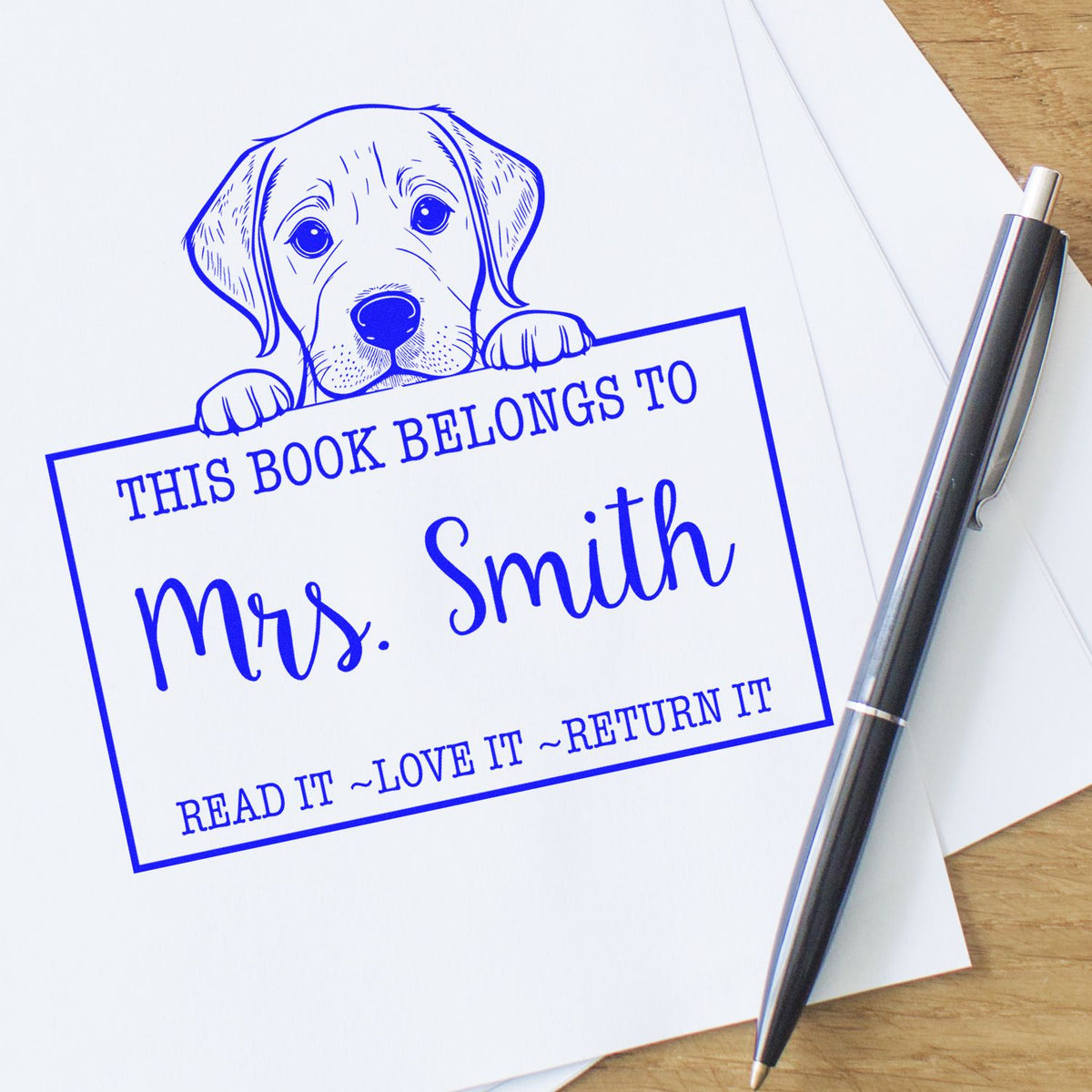 Blue self-inking book stamp and seal with puppy design, Mrs. Smith text, on white paper beside a silver pen